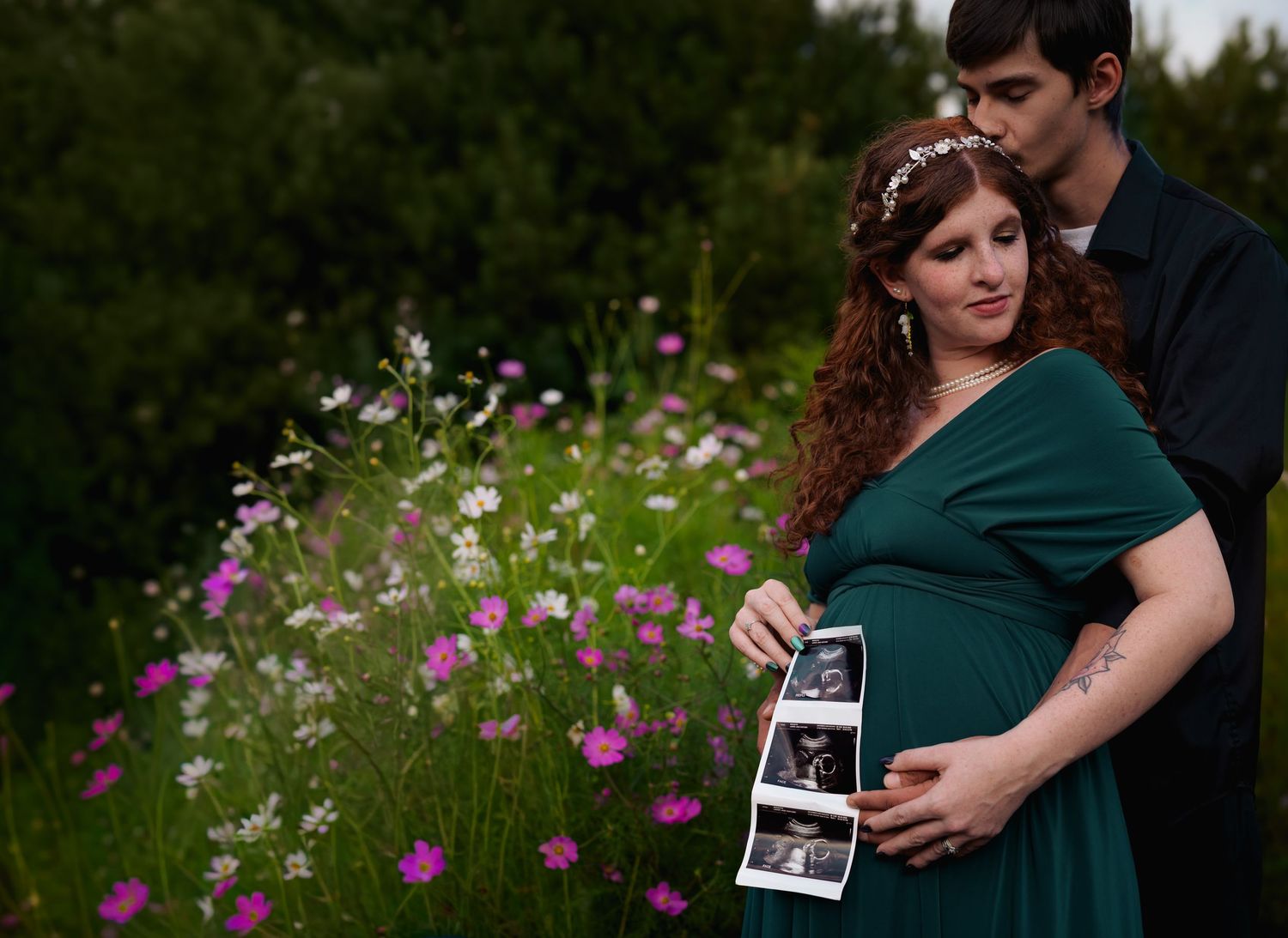 Pregnant woman in green dress holds ultrasound photos with partner among wildflowers in a maternity announcement outdoor shoot.
