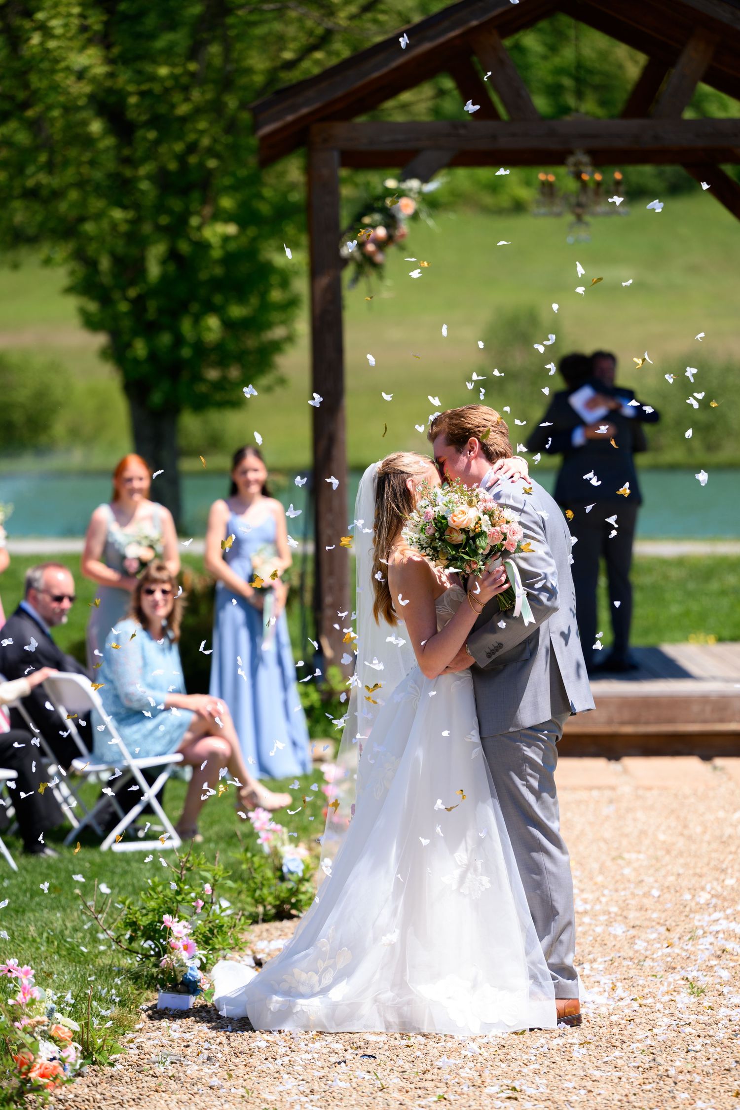 Bride and groom share first kiss outdoors under wooden arbor as confetti falls during sunny wedding ceremony.