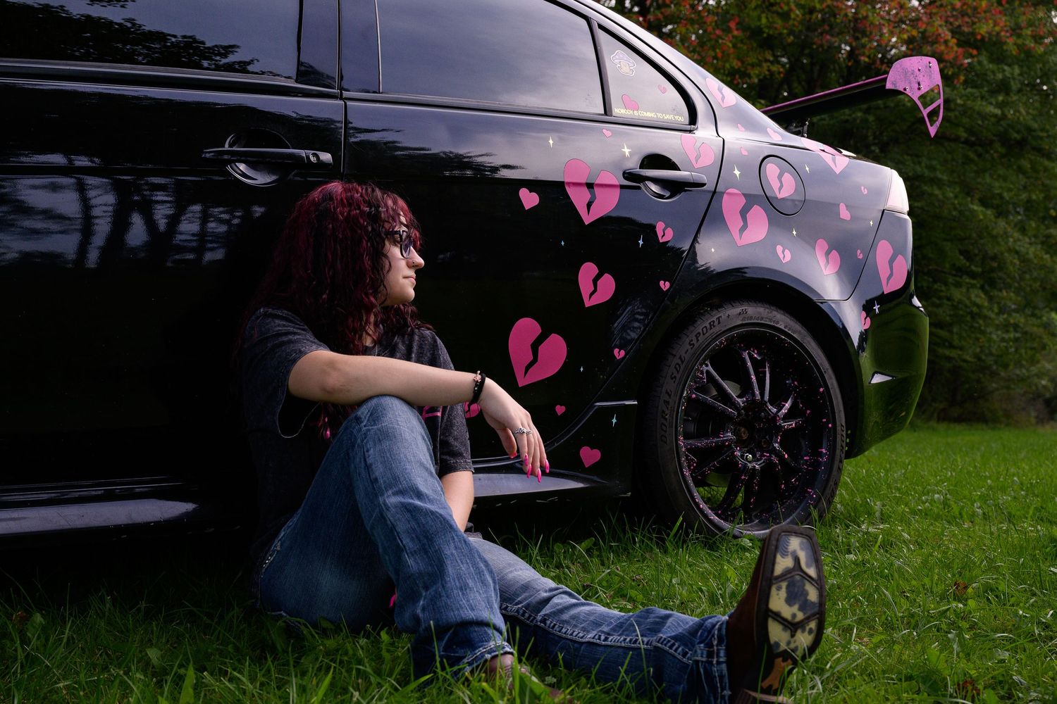 Person sitting beside a black car decorated with pink broken heart designs on green grass.
