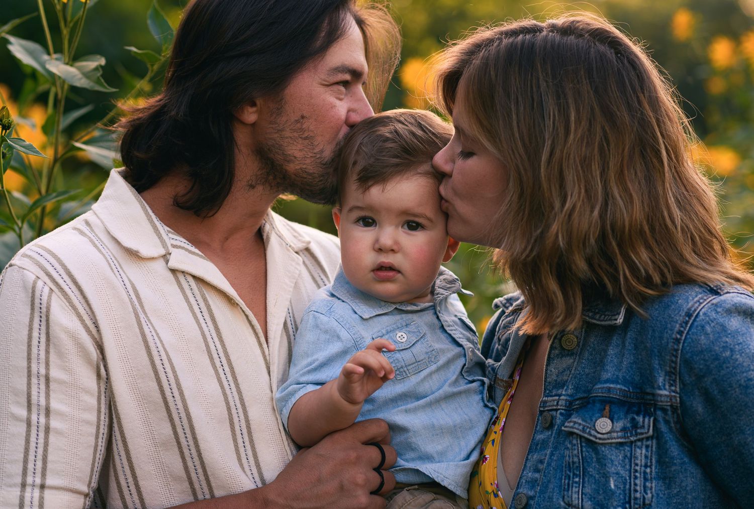 A couple kisses their baby boy on the cheeks in a sunny flower field, capturing a warm family moment.