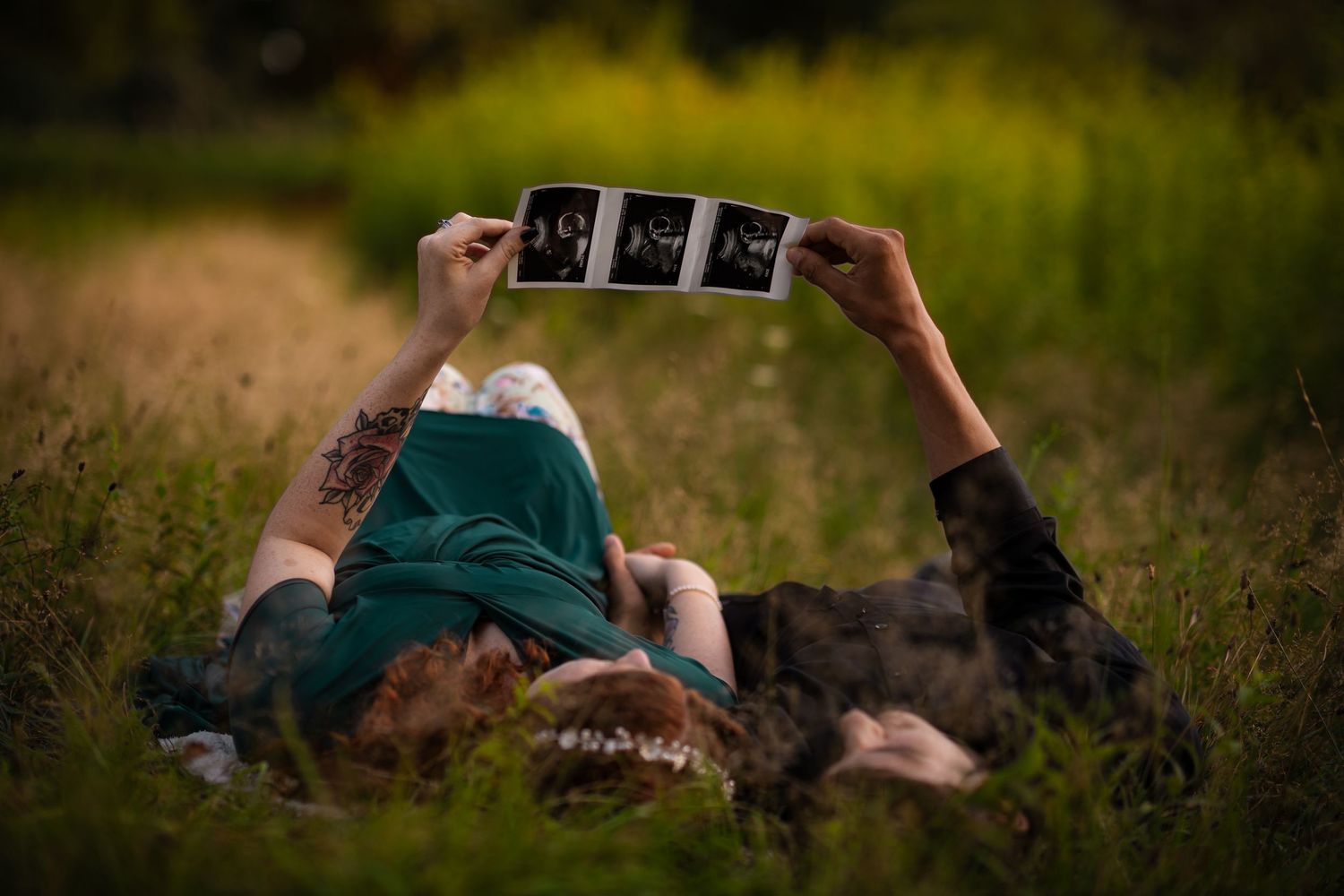 Pregnant woman lying in grass holding ultrasound photos above her with a dog beside her.