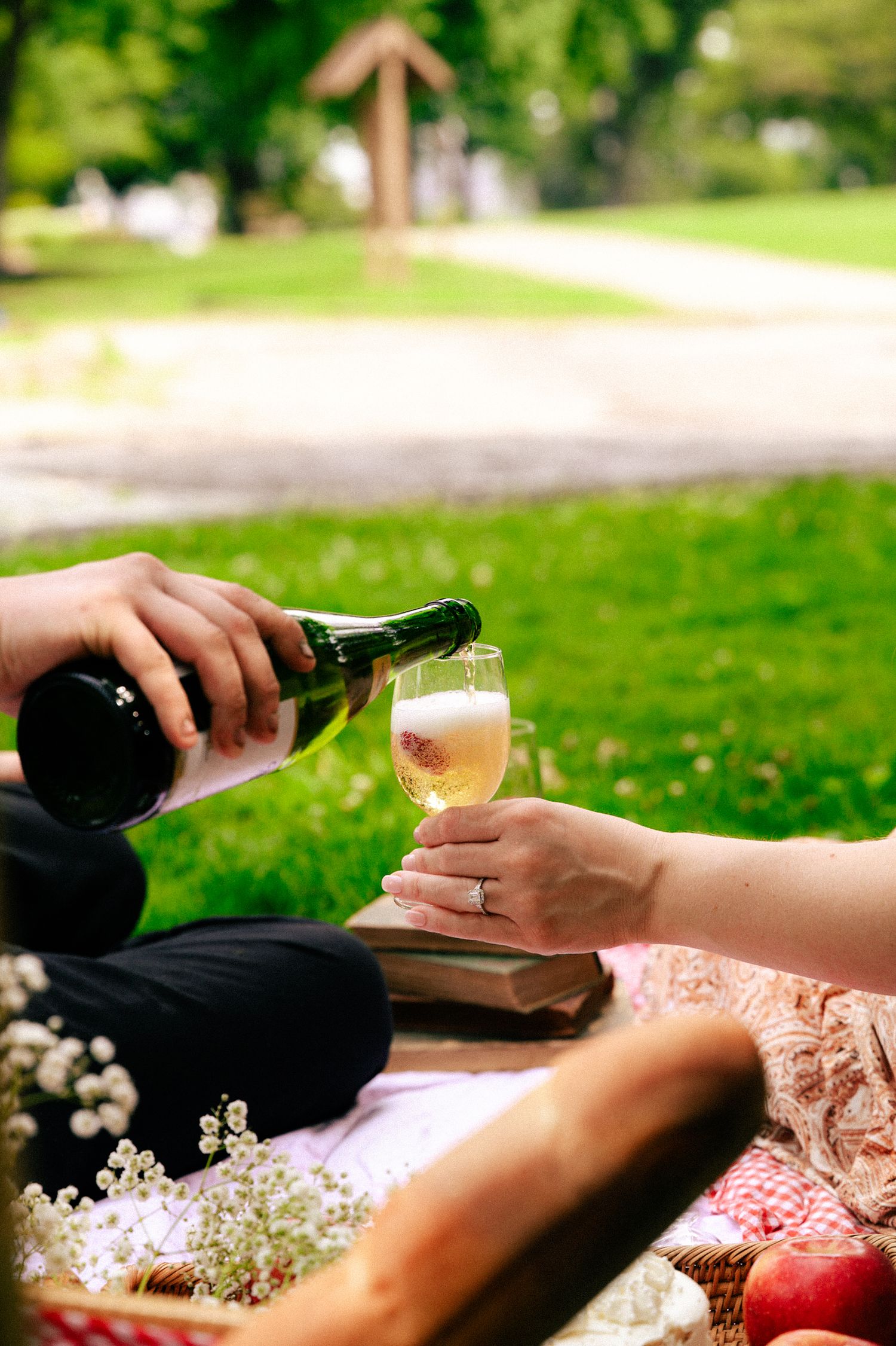 Person pouring champagne into a glass during an outdoor picnic on green grass with flowers and fruit nearby.