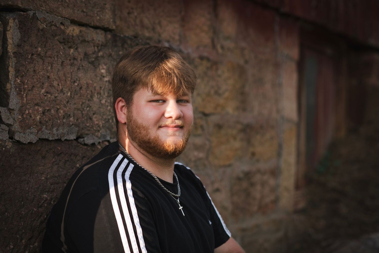 Young man with red beard smiling, wearing black Adidas shirt, leaning against rustic brick wall outdoors.