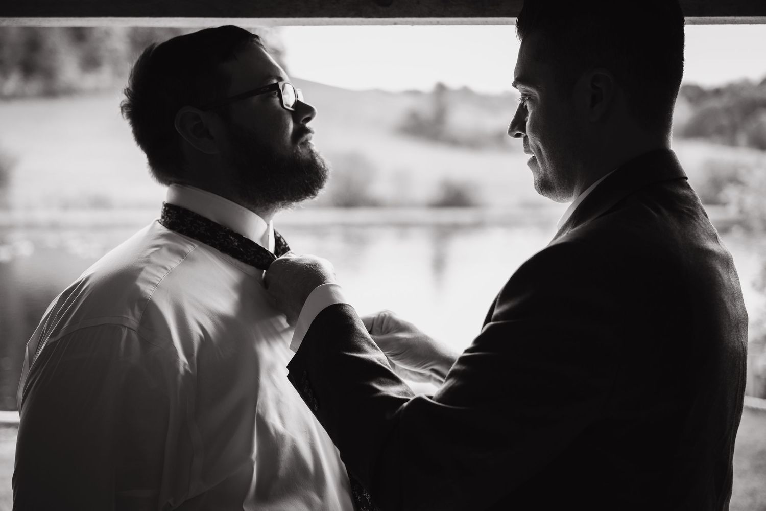 Black and white photo of groomsman adjusting grooms bow tie before wedding ceremony near window.