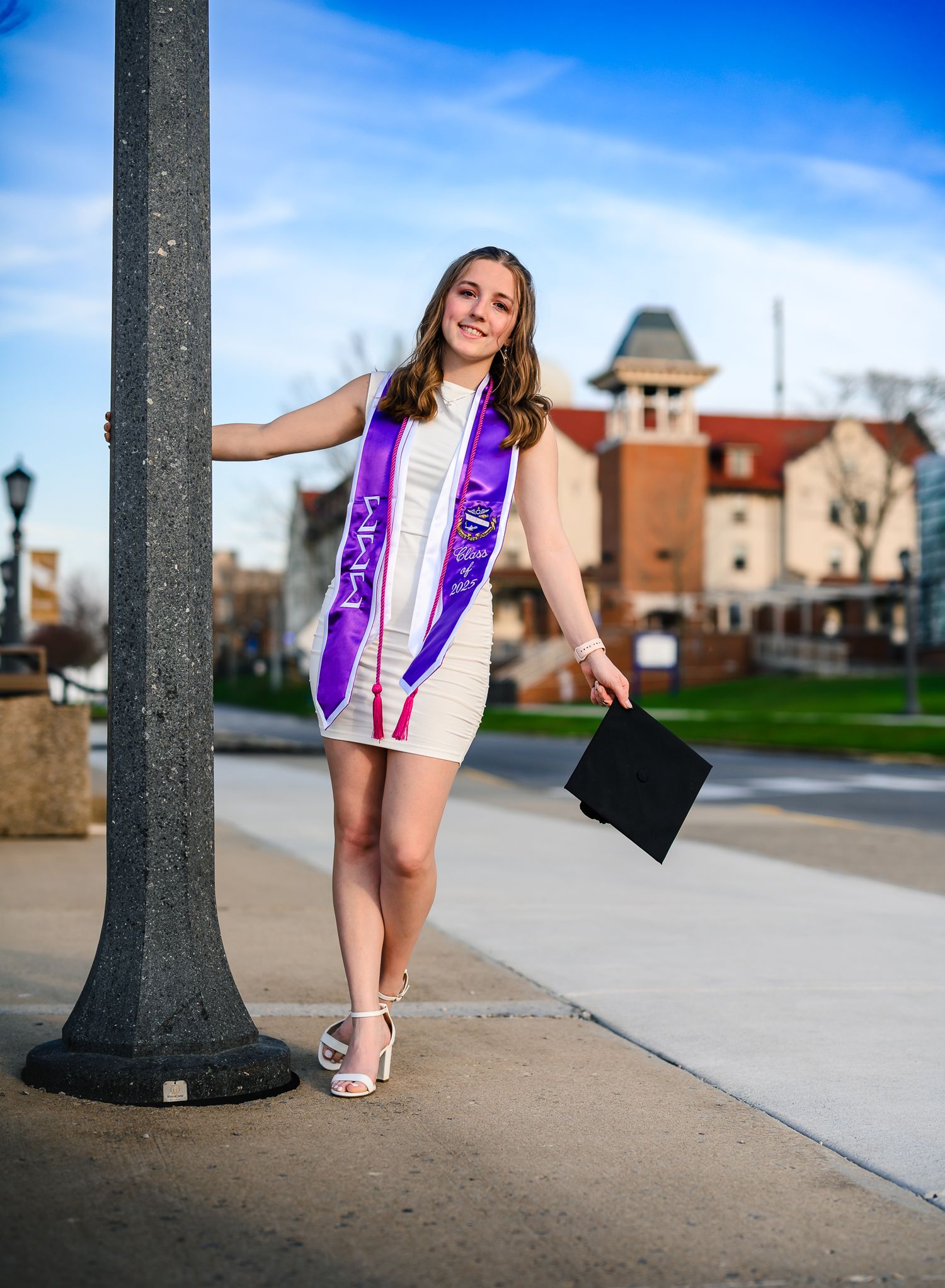 Smiling graduate poses with cap and purple sash by lamppost on sunny campus walkway in white dress.