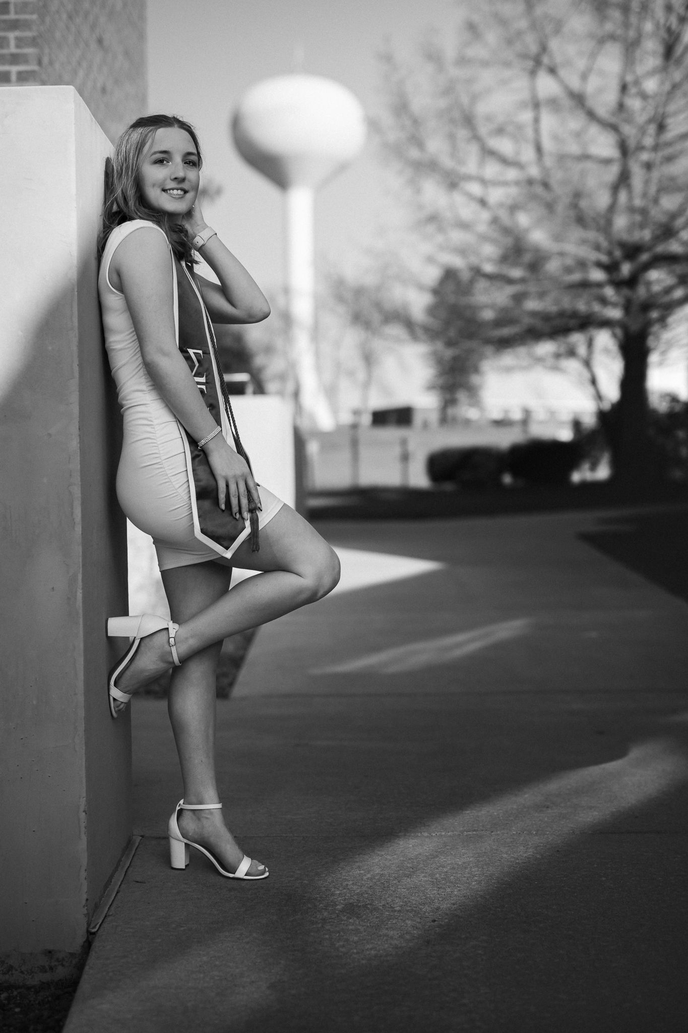 Black and white photo of a smiling young woman in a short dress and heels leaning against a wall outdoors.