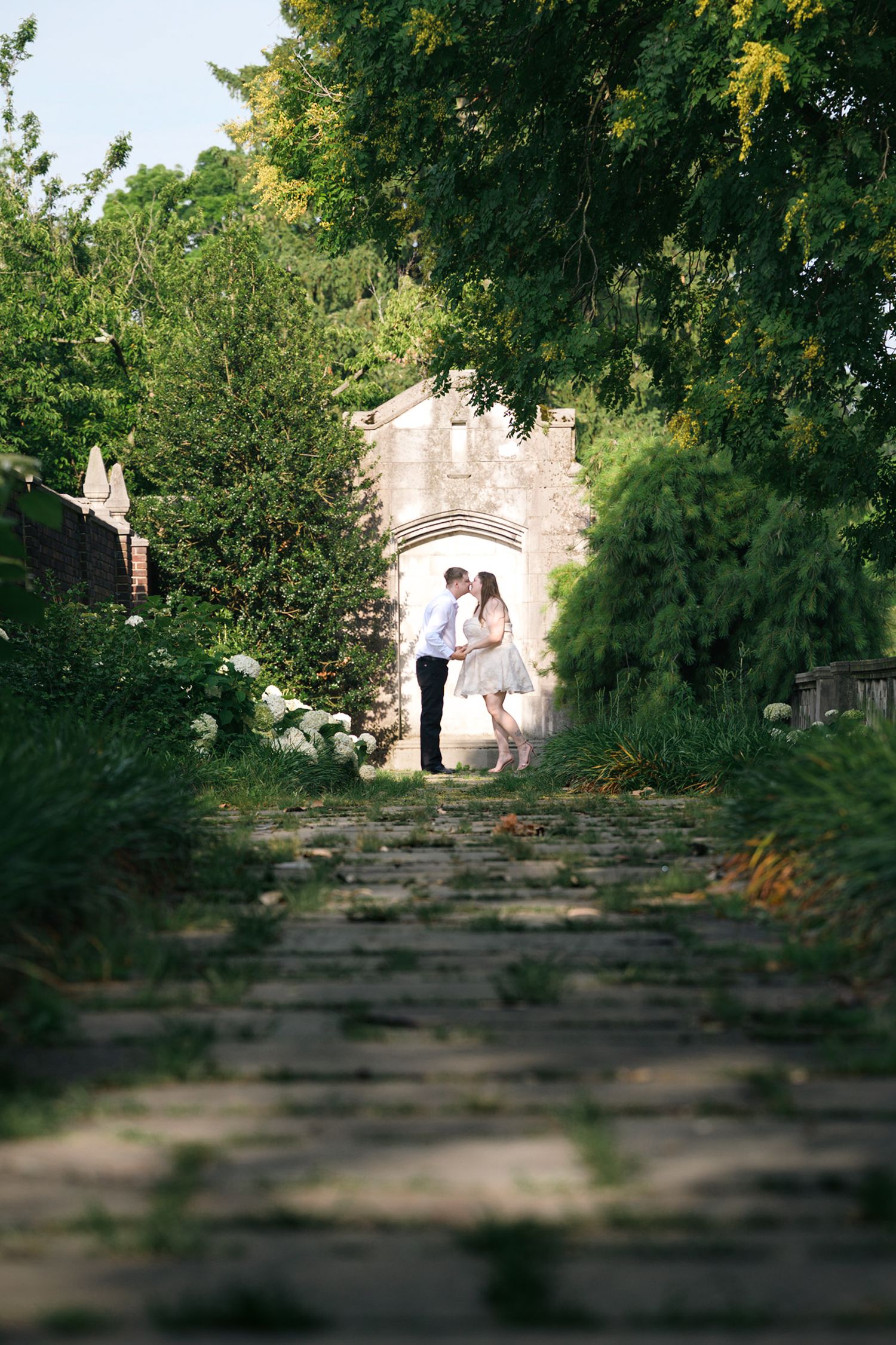 Couple sharing a romantic kiss on a cobblestone path surrounded by lush green trees leading to a stone archway.