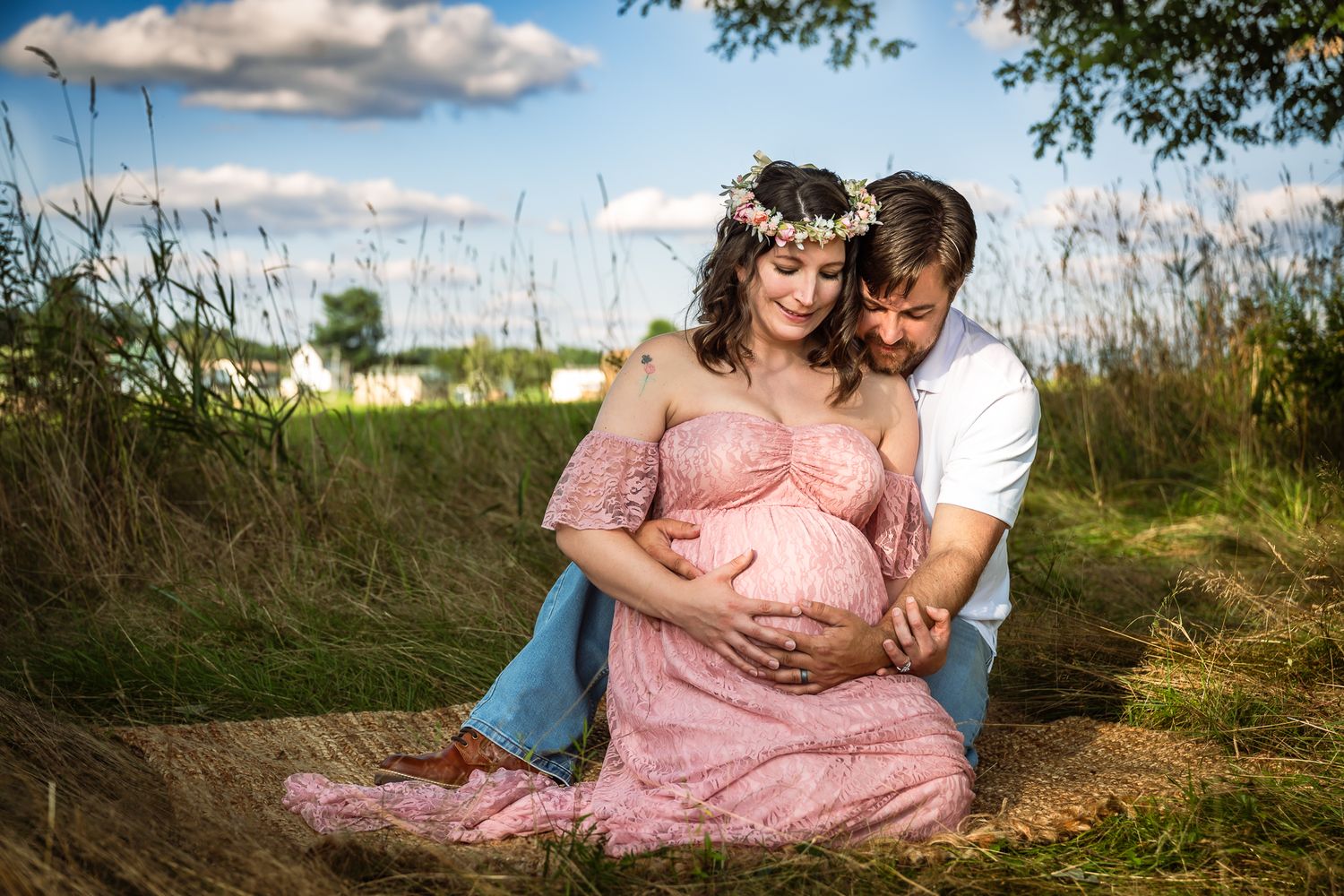 Couple sitting outdoors during maternity shoot, pregnant woman in pink lace dress leaning on partner.