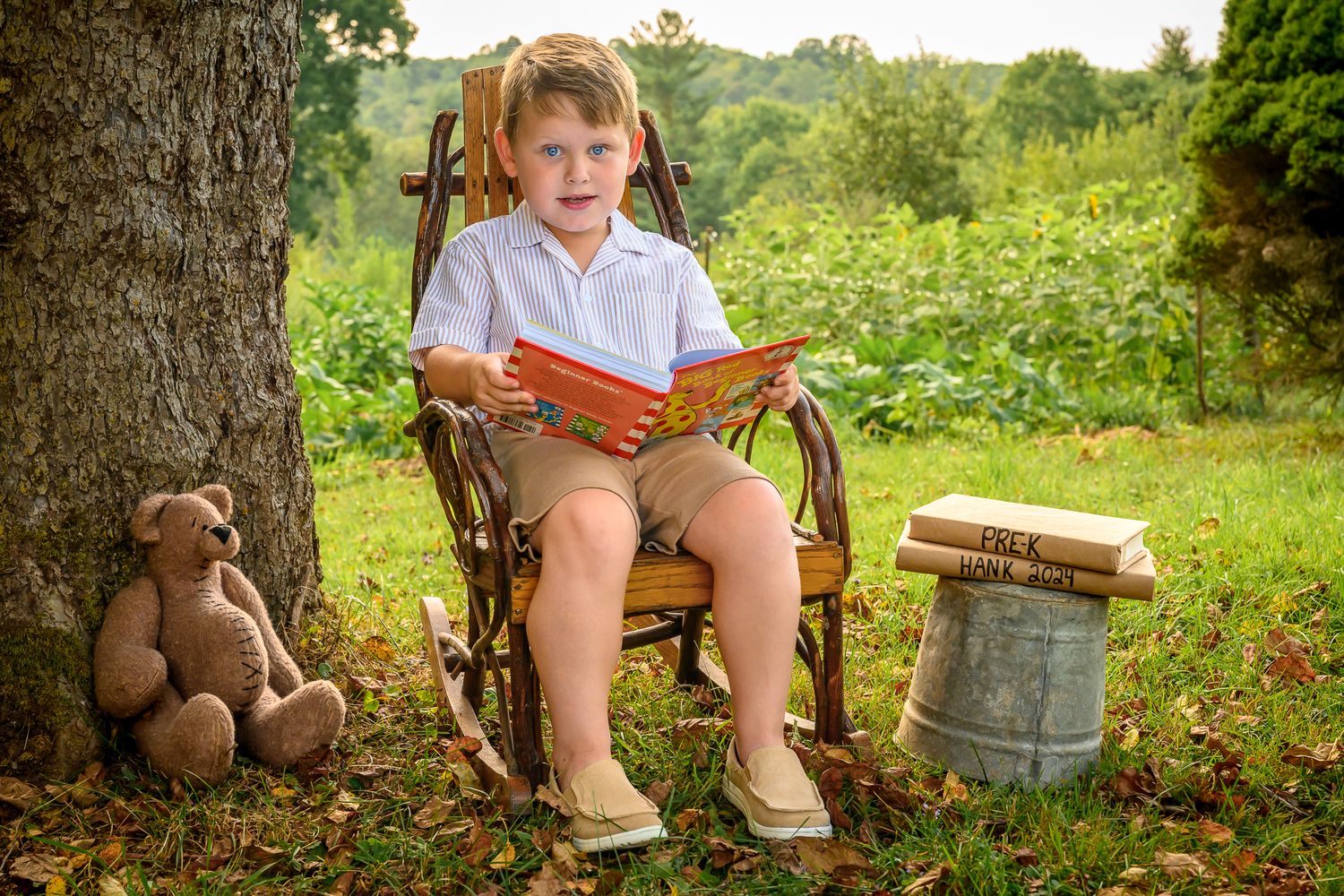 Young boy reading a book in a rustic rocking chair outdoors, with a teddy bear beside him in a lush green garden.