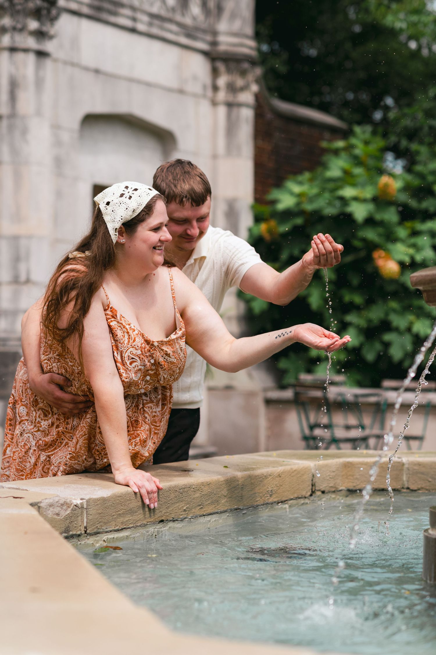 A smiling couple plays with water at a stone fountain outdoors, surrounded by lush green gardens.