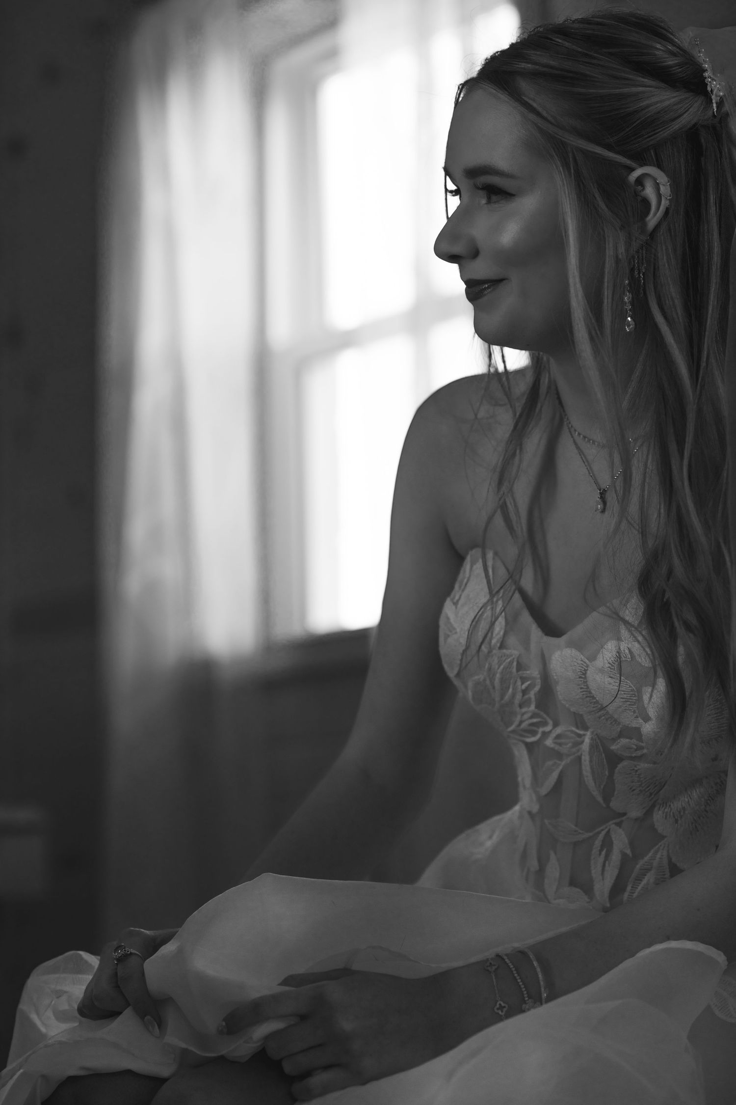 Black and white portrait of a smiling bride in a wedding dress sitting by a window with soft natural light.
