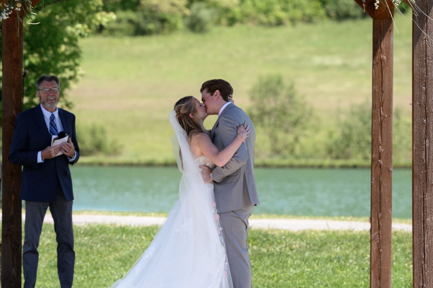 Bride and groom share first kiss at outdoor wedding ceremony near a pond, officiant smiling beside them.