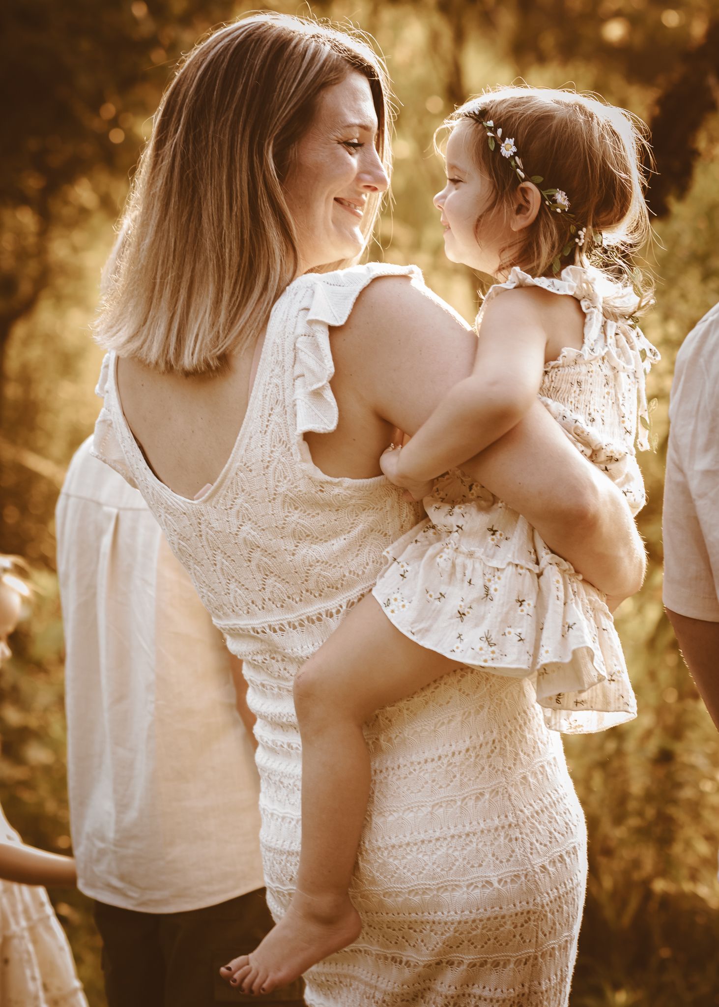 A mother in a white lace dress holds her toddler daughter in a sunlit outdoor portrait.