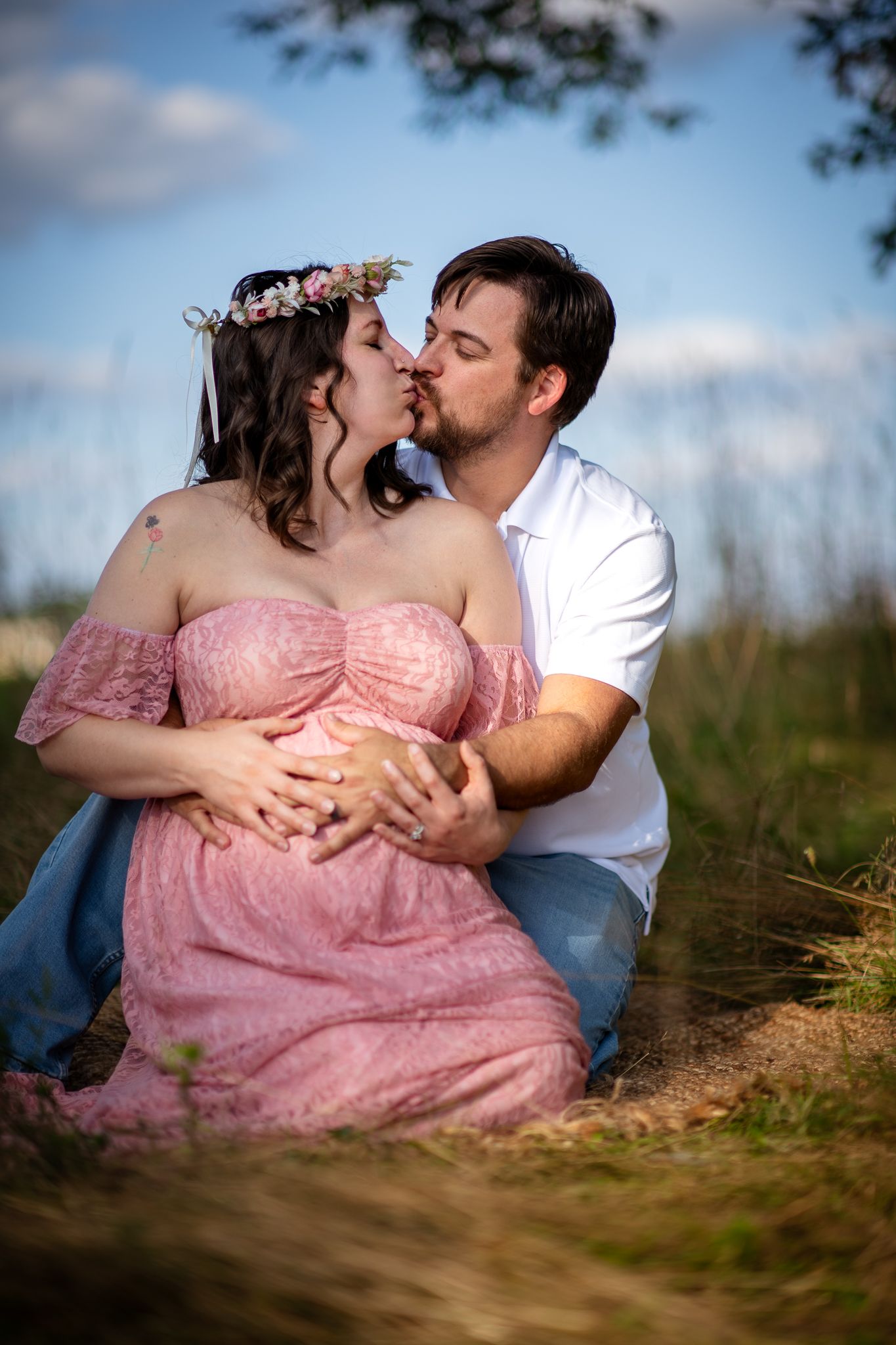 Couple sharing a kiss during outdoor maternity photo shoot, pregnant woman in pink lace dress with floral crown.