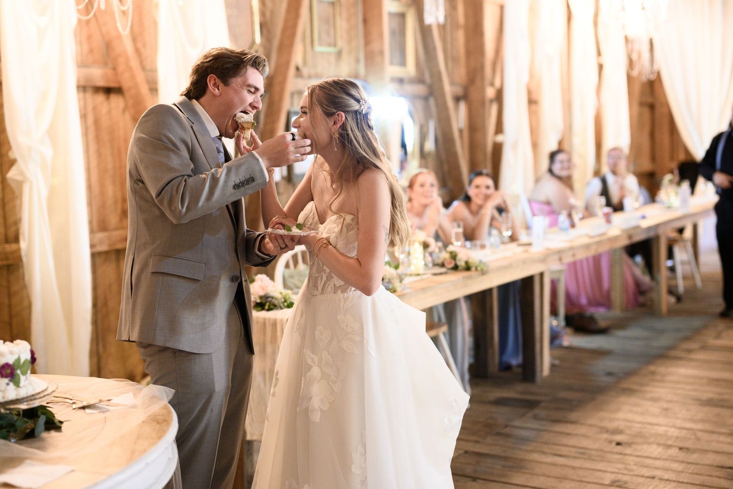 Bride and groom share cake during rustic barn wedding reception with guests seated at long decorated tables.
