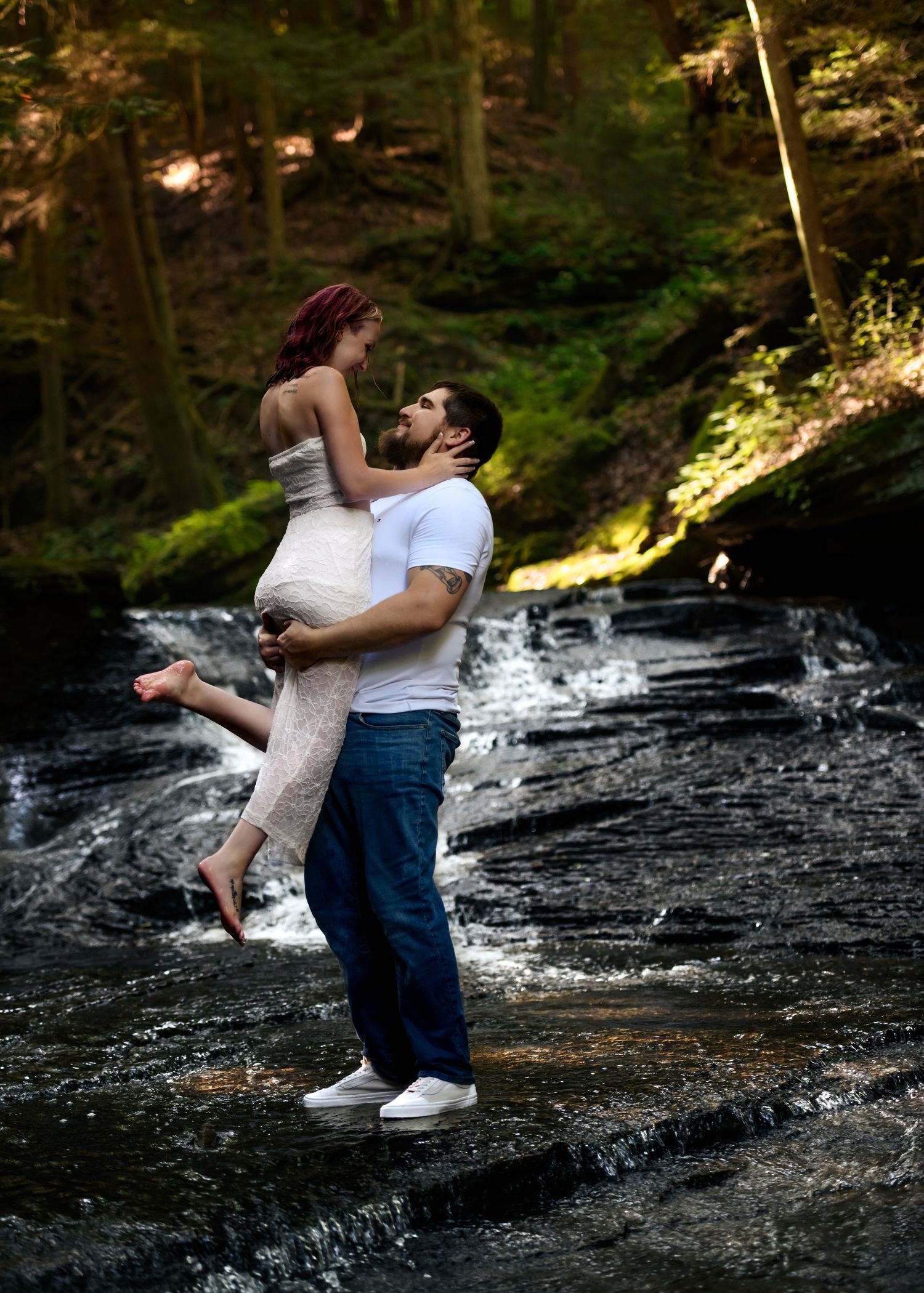 Couple sharing a romantic moment by a forest waterfall, man lifting woman in white dress near rocky stream.