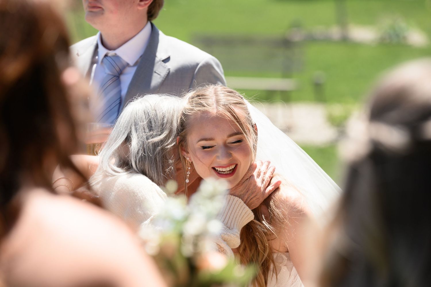 A smiling bride in a white veil shares a warm hug with an older woman at an outdoor wedding ceremony.