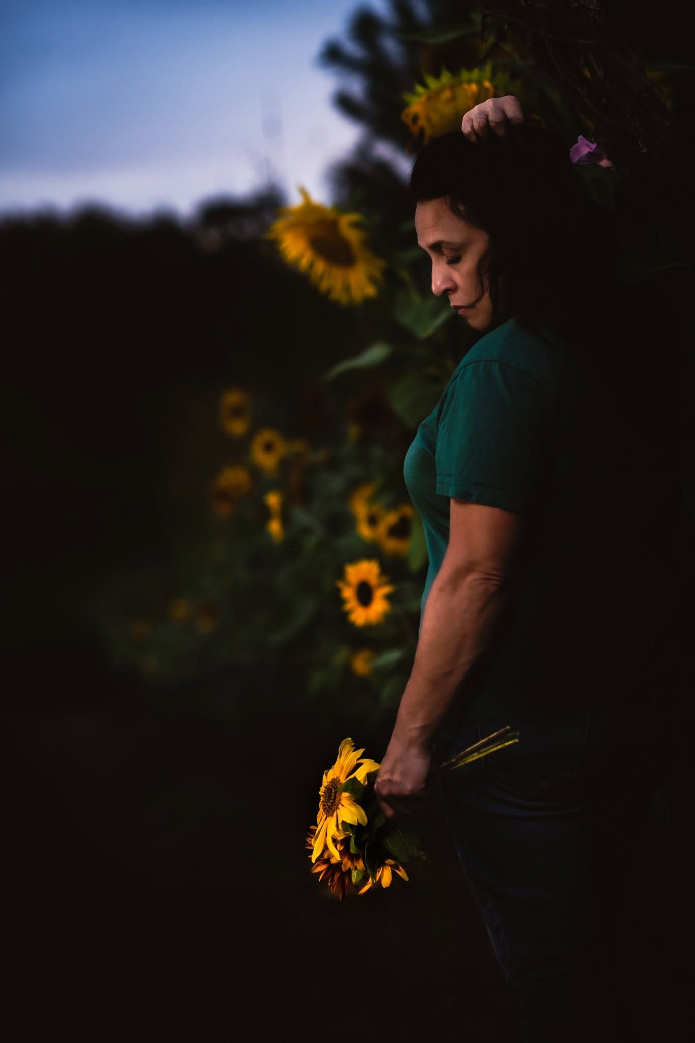 Woman in green shirt holds sunflowers at dusk, standing near a sunflower field in soft evening light.