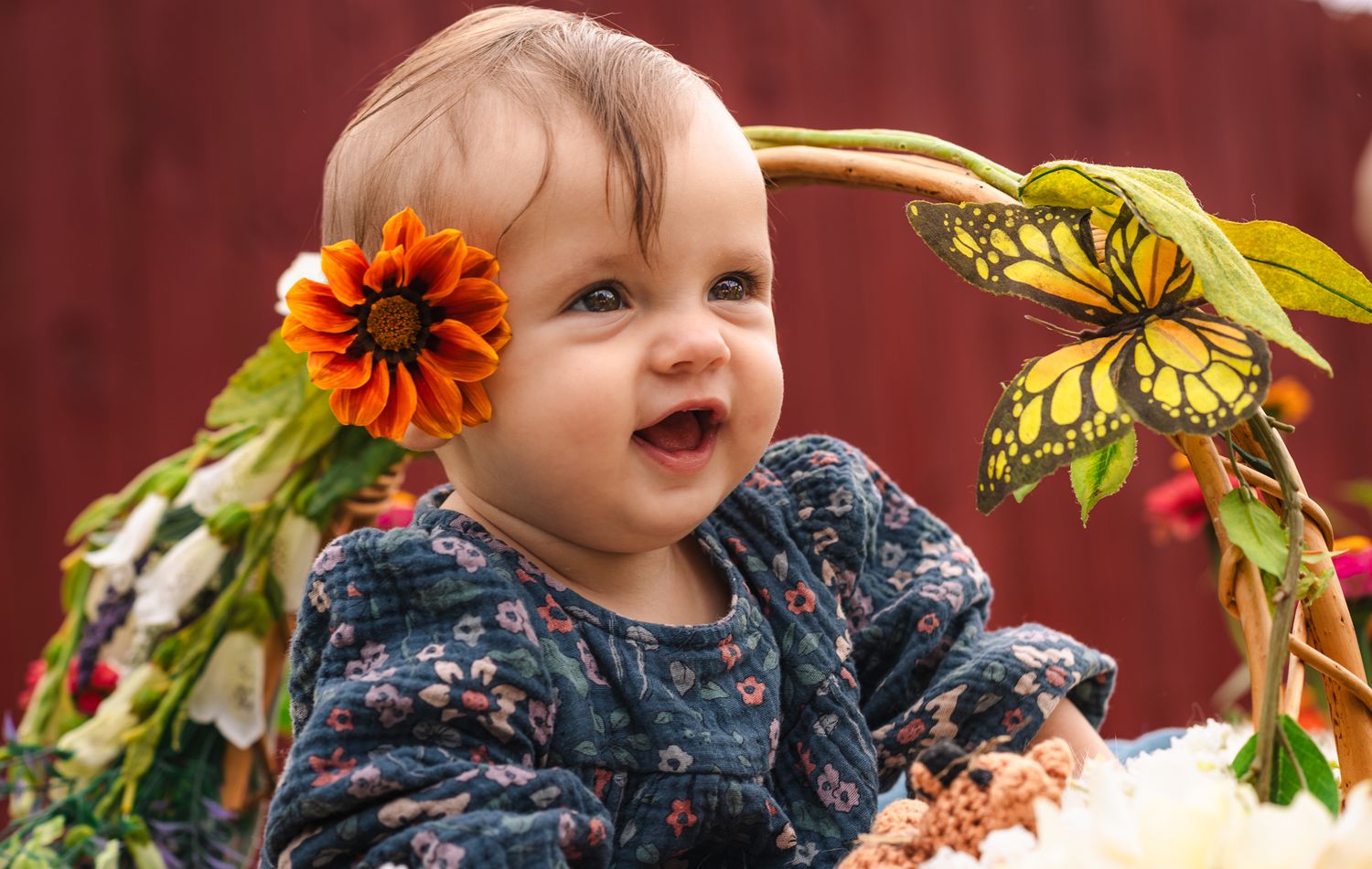 Smiling baby girl with orange flower in hair sits among colorful flowers and butterfly decorations outdoors.