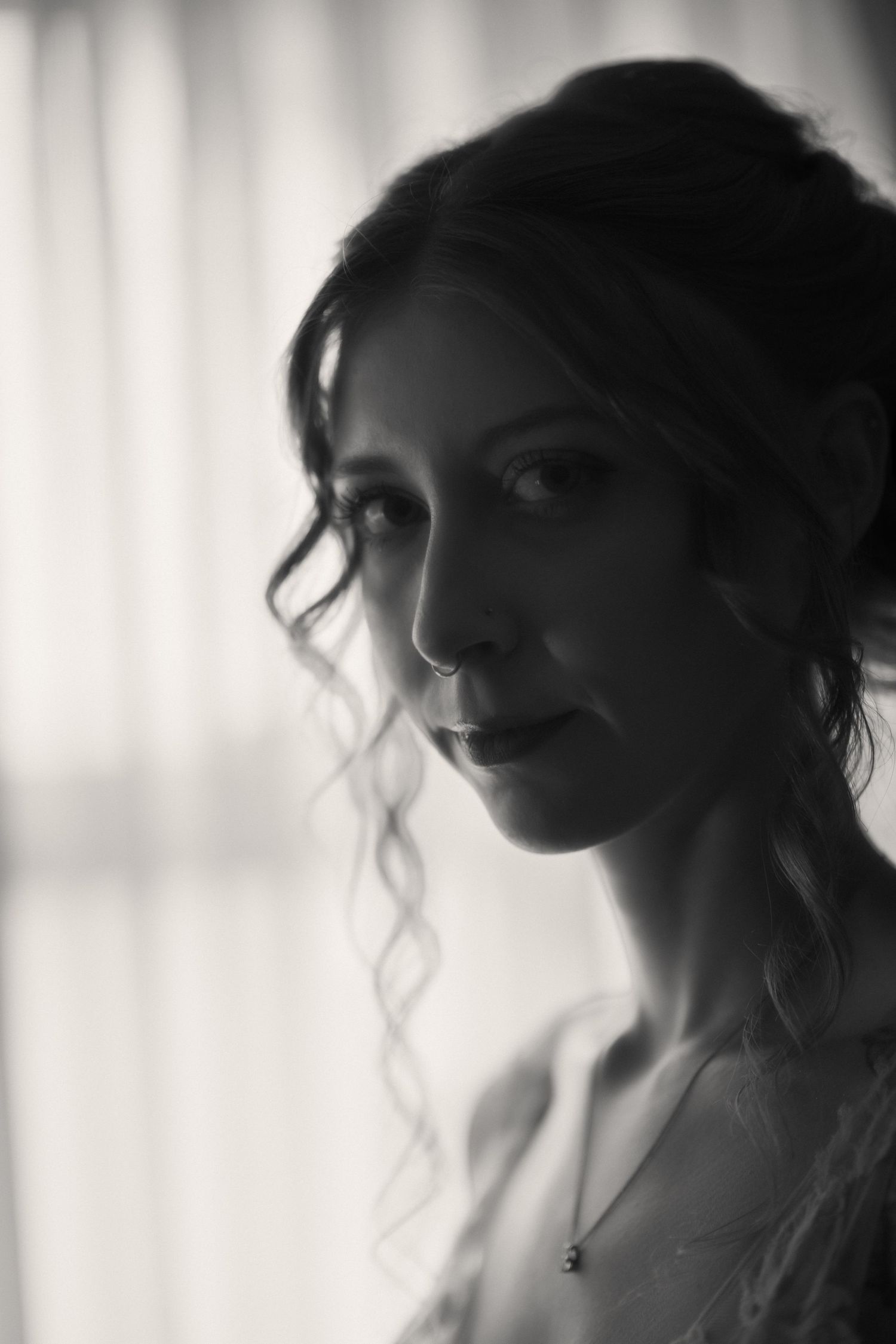 Black and white portrait of a young woman with curly updo hair, wearing a necklace, posed near a softly lit window.