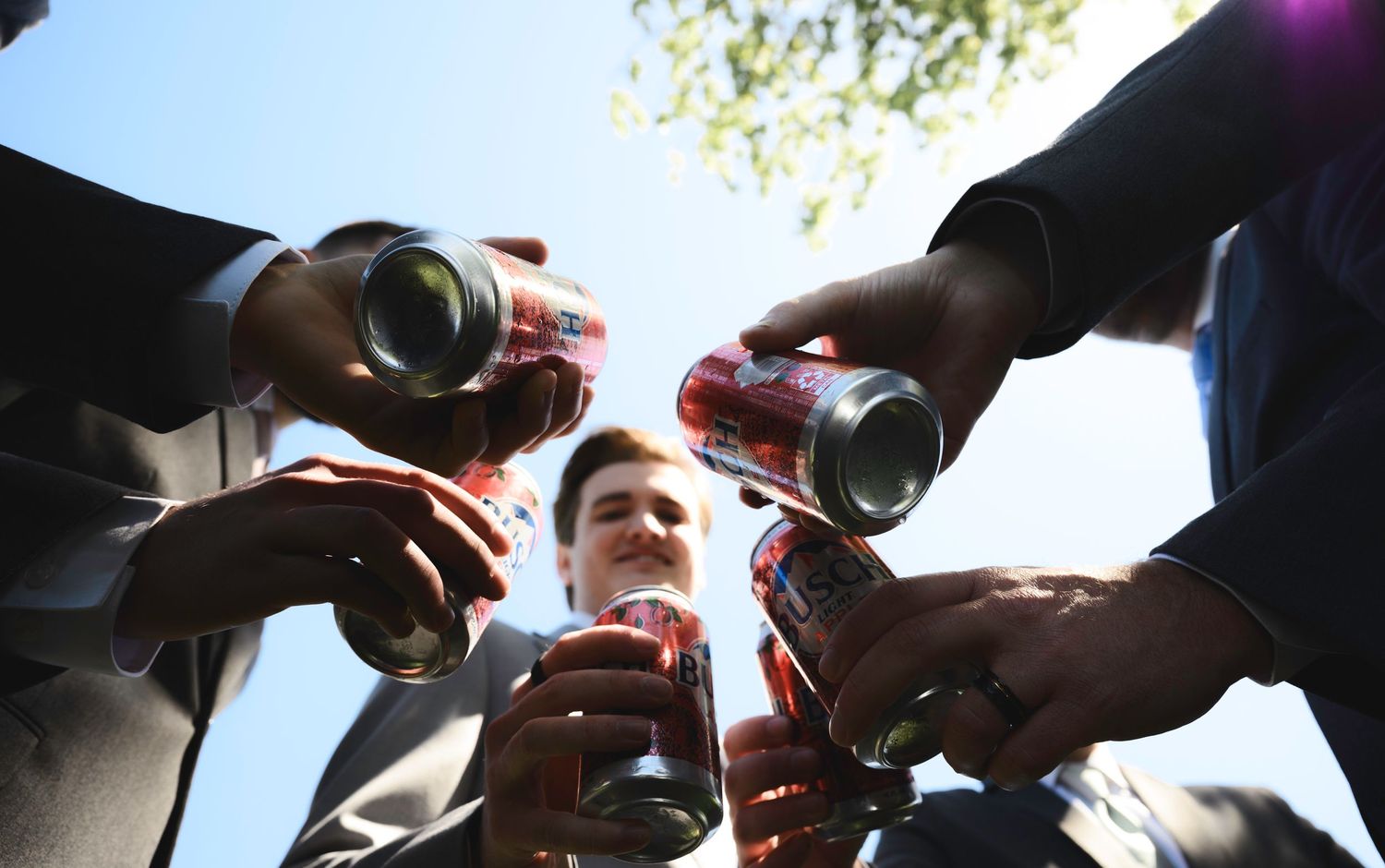 Group of men in suits toasting with red cans of soda outdoors, viewed from below against a bright sky.