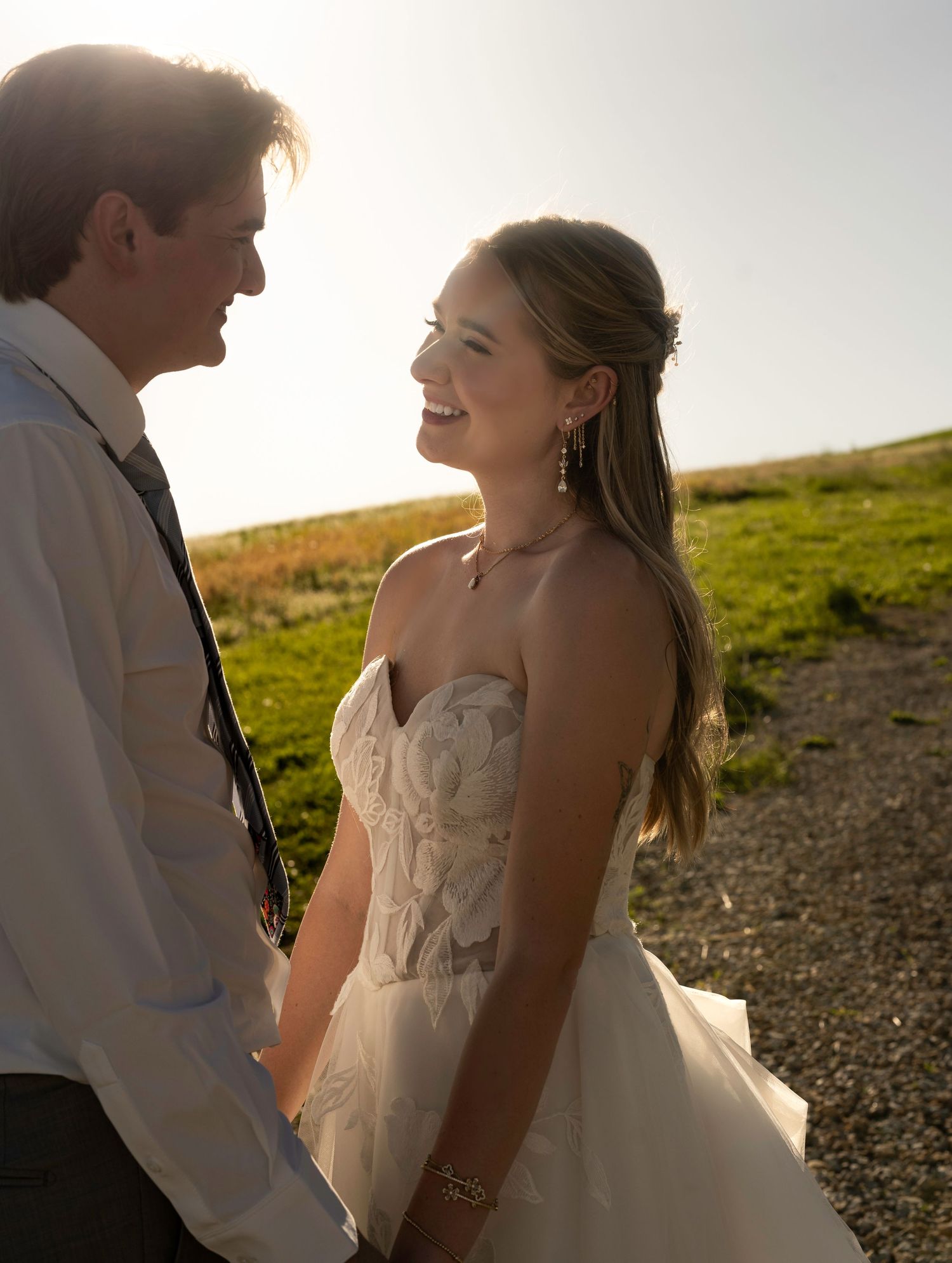 A bride in a strapless floral gown smiles at her groom while holding hands outdoors in golden hour light.