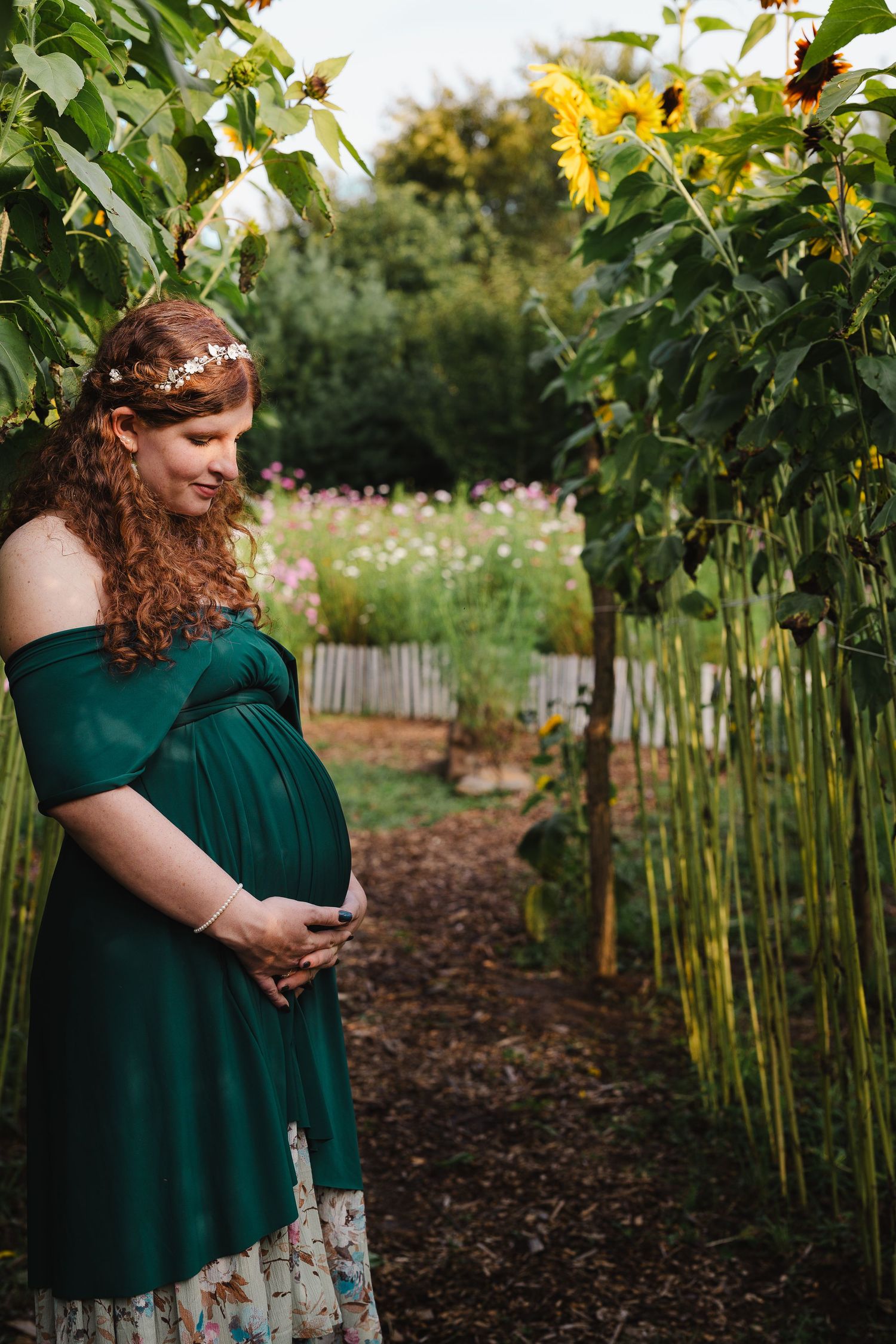 Pregnant woman in green off-shoulder dress holding belly, standing in sunflower garden path wearing floral crown.
