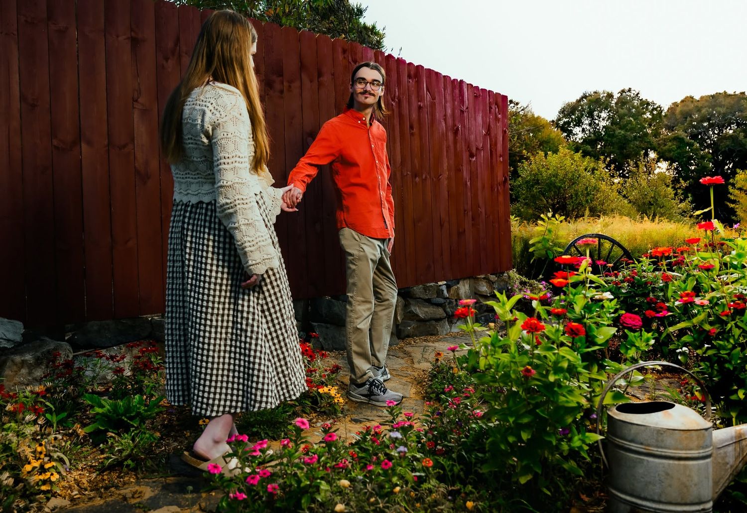 Couple holding hands in a colorful garden with red flowers near a dark wooden fence.