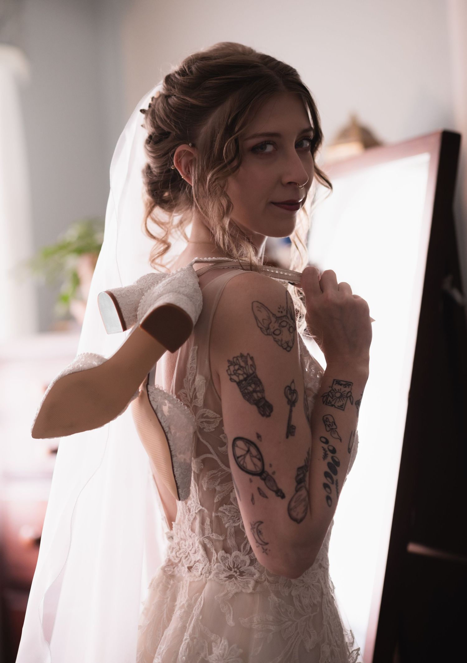 Tattooed bride in lace wedding dress and veil poses elegantly in front of mirror on her wedding day.