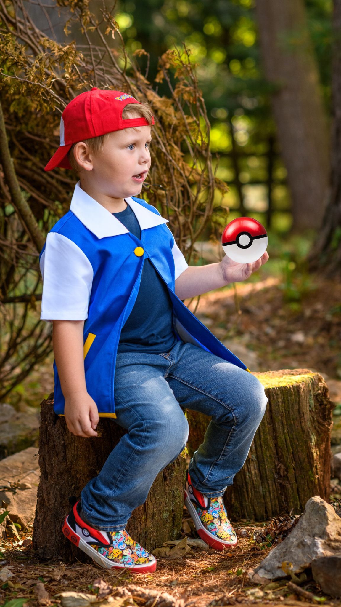 A young boy dressed as Ash Ketchum from Pokemon holds a Pokeball while sitting on a tree stump outdoors.
