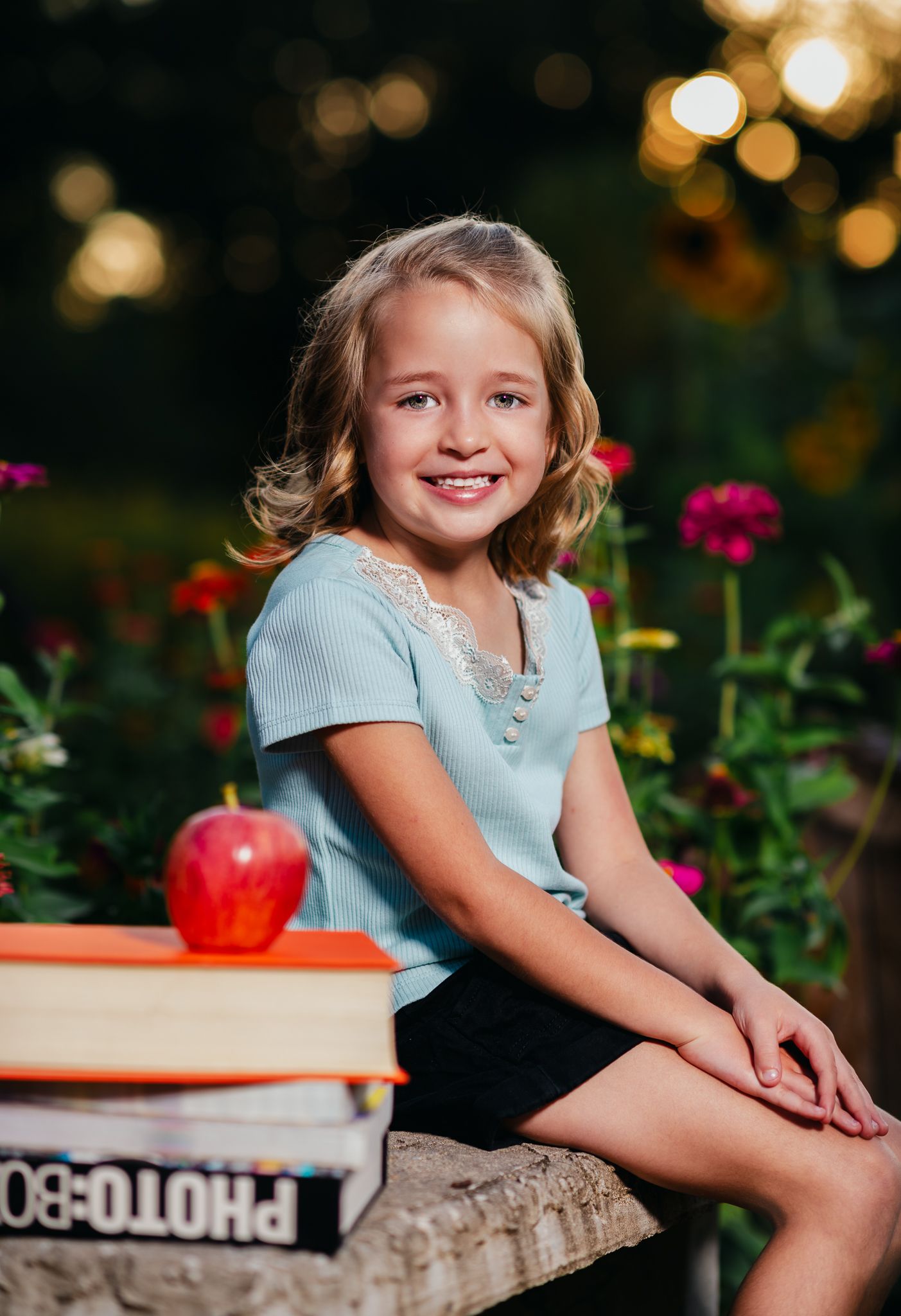 Smiling young girl in blue top sits outdoors with stacked books and red apple, surrounded by colorful flowers.