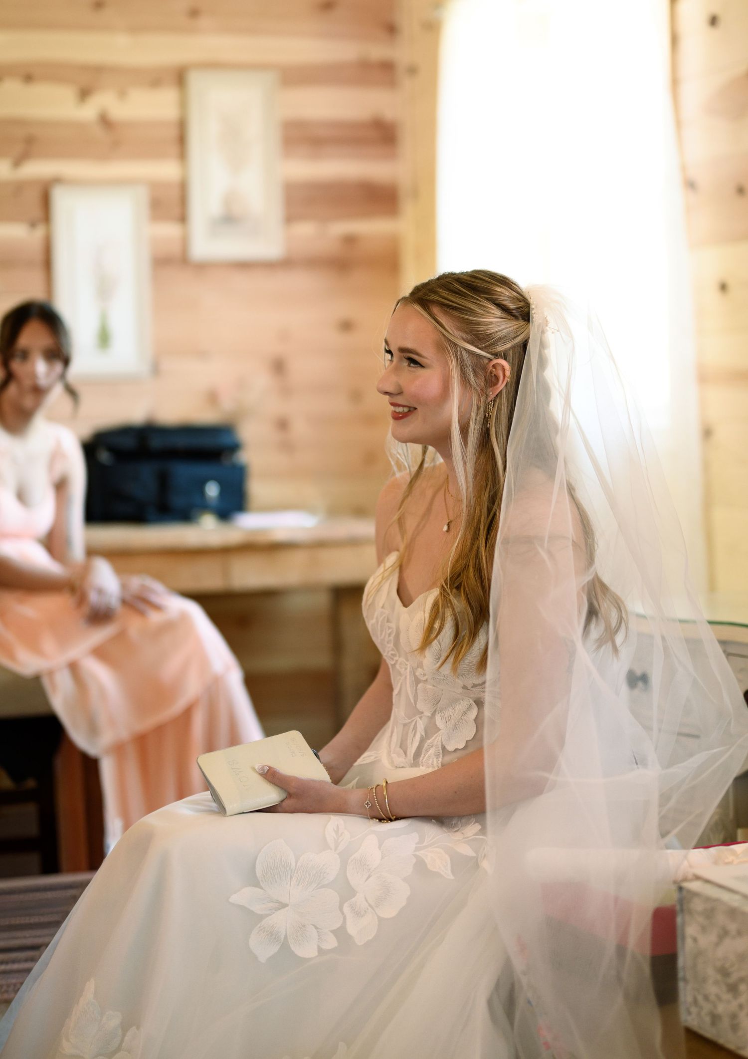 Smiling bride in white lace gown and veil sits holding a book in a wooden cabin room with bridesmaid in background.