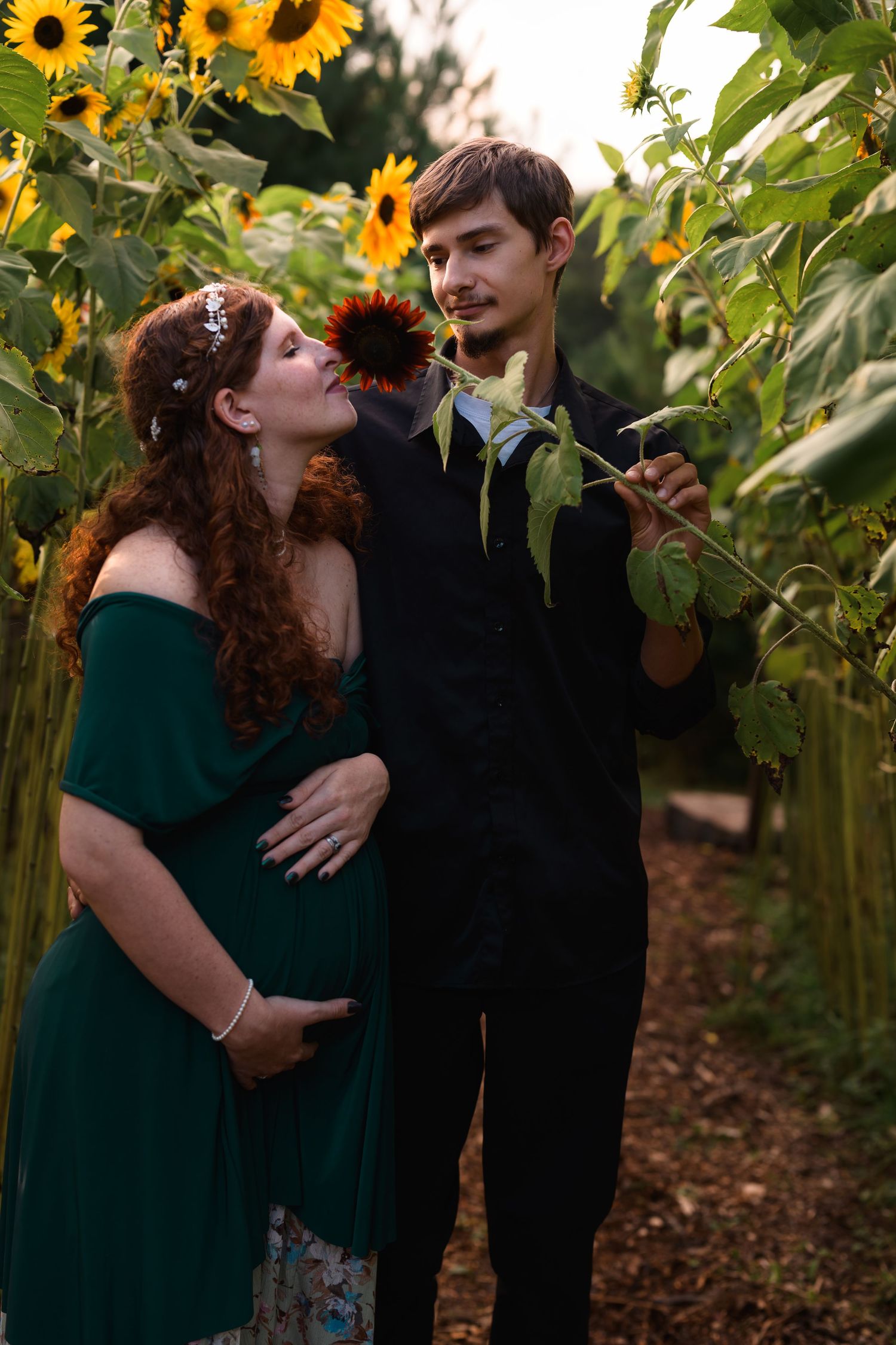Pregnant woman in teal dress with partner holding sunflower in a sunflower field during maternity shoot.