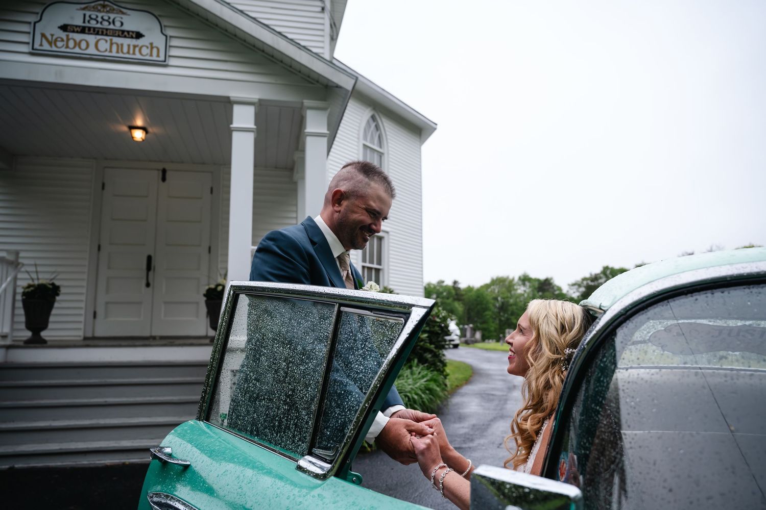 Groom helping bride into a vintage green car outside Mount Nebo Church on their wedding day.