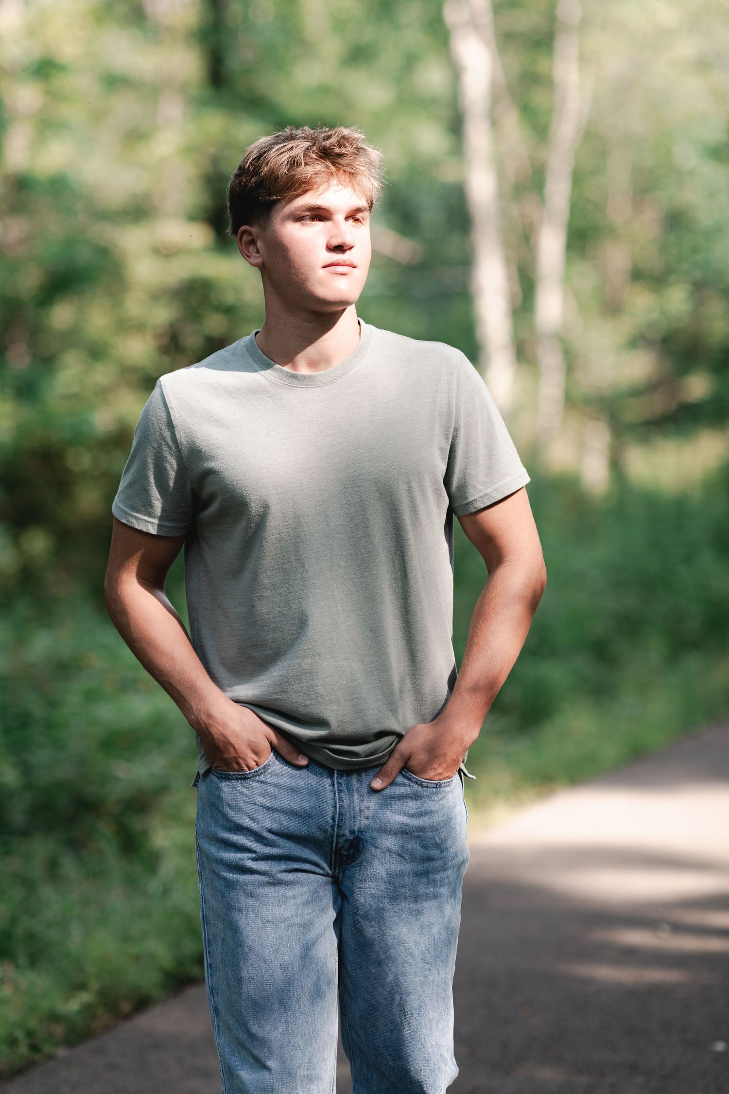 Young man in gray t-shirt and jeans standing on a path in a lush green forest, hands in pockets.