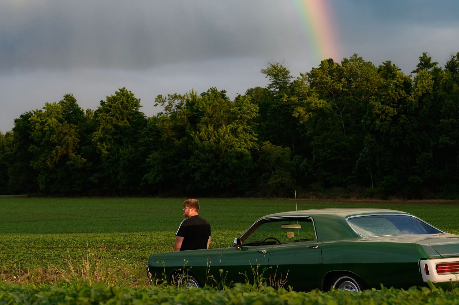 Man standing beside a green classic muscle car in a field with a rainbow over lush trees in the background.