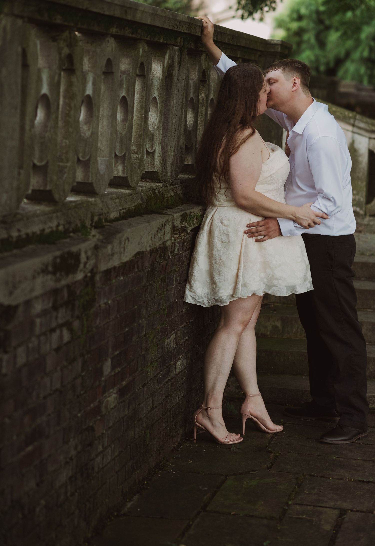 Couple sharing a romantic kiss against a stone balustrade wall, woman in white dress and heels, man in white shirt.