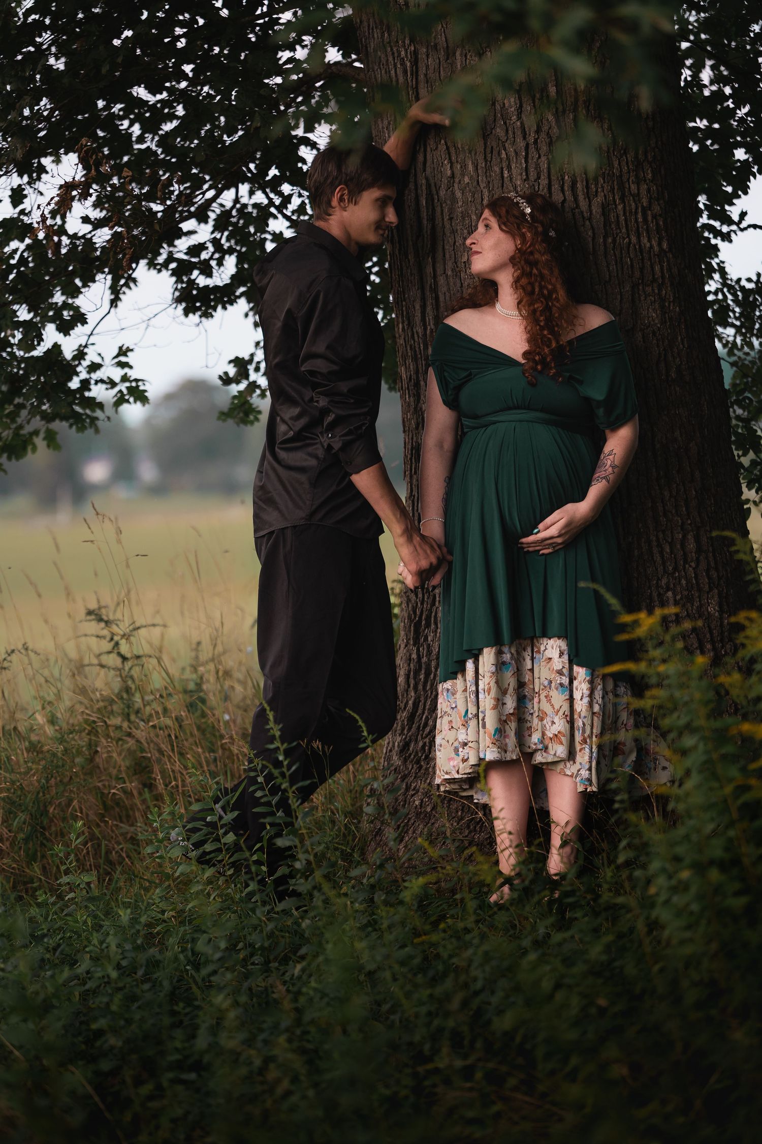 Pregnant woman in green dress holds hands with partner by a tree in a lush outdoor field setting.