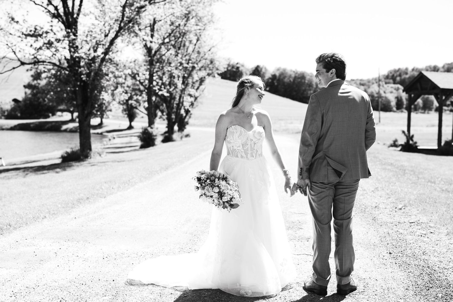 Black and white photo of a bride and groom holding hands walking down a sunny tree-lined path on their wedding day.