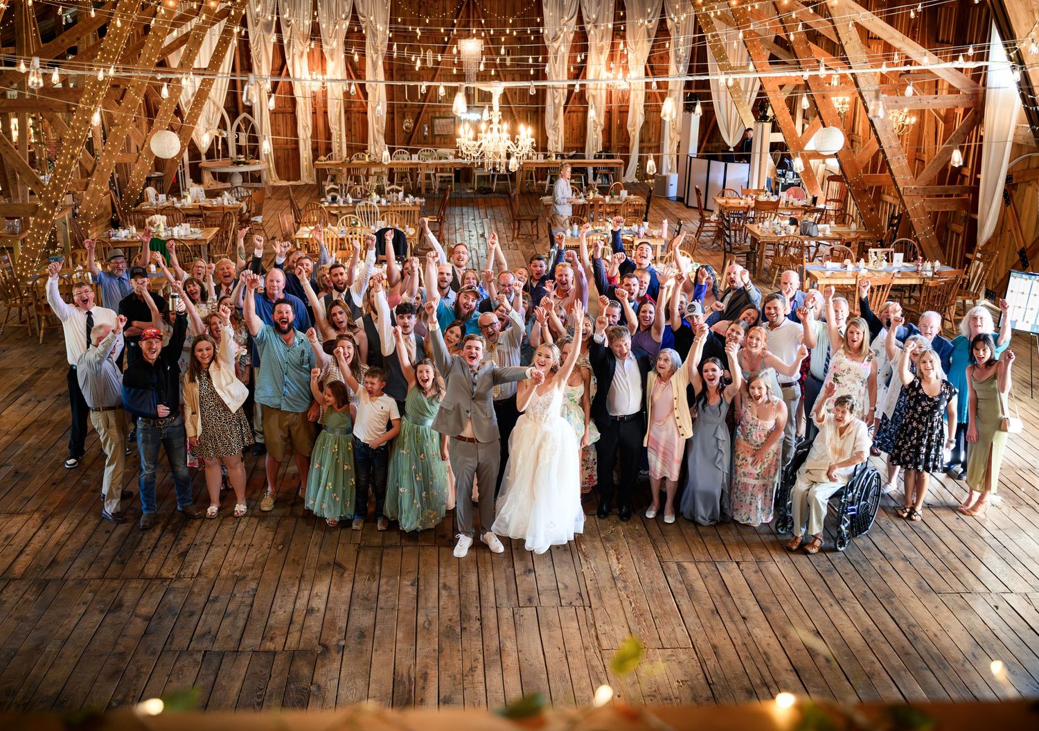 Large wedding group photo inside a rustic barn venue with guests cheering and raising hands in celebration.