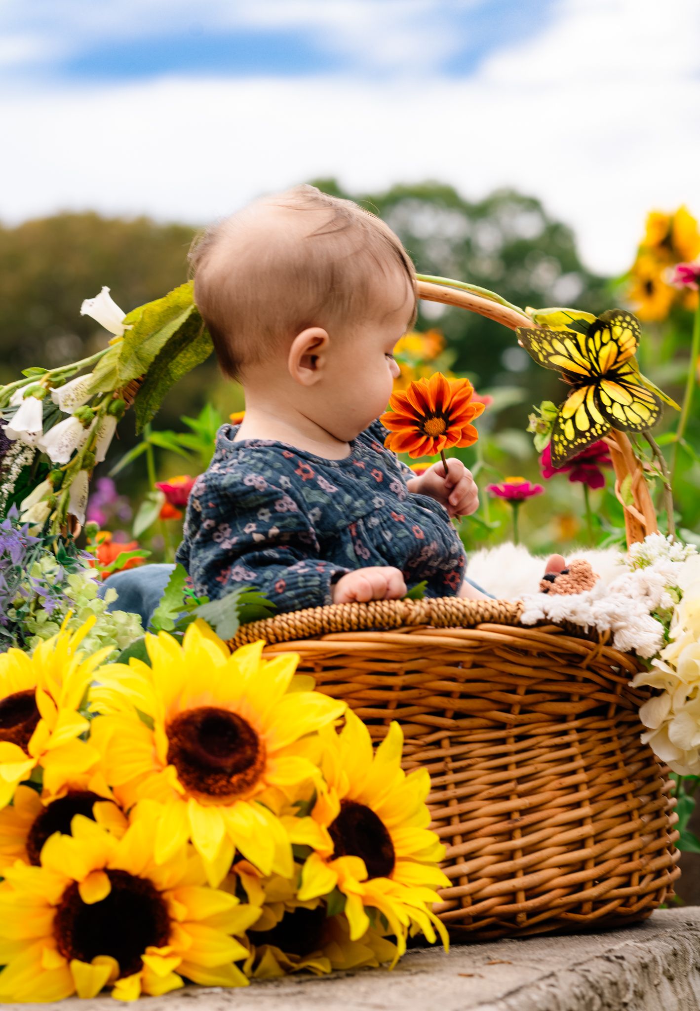 Baby sitting in wicker basket surrounded by colorful flowers and sunflowers.