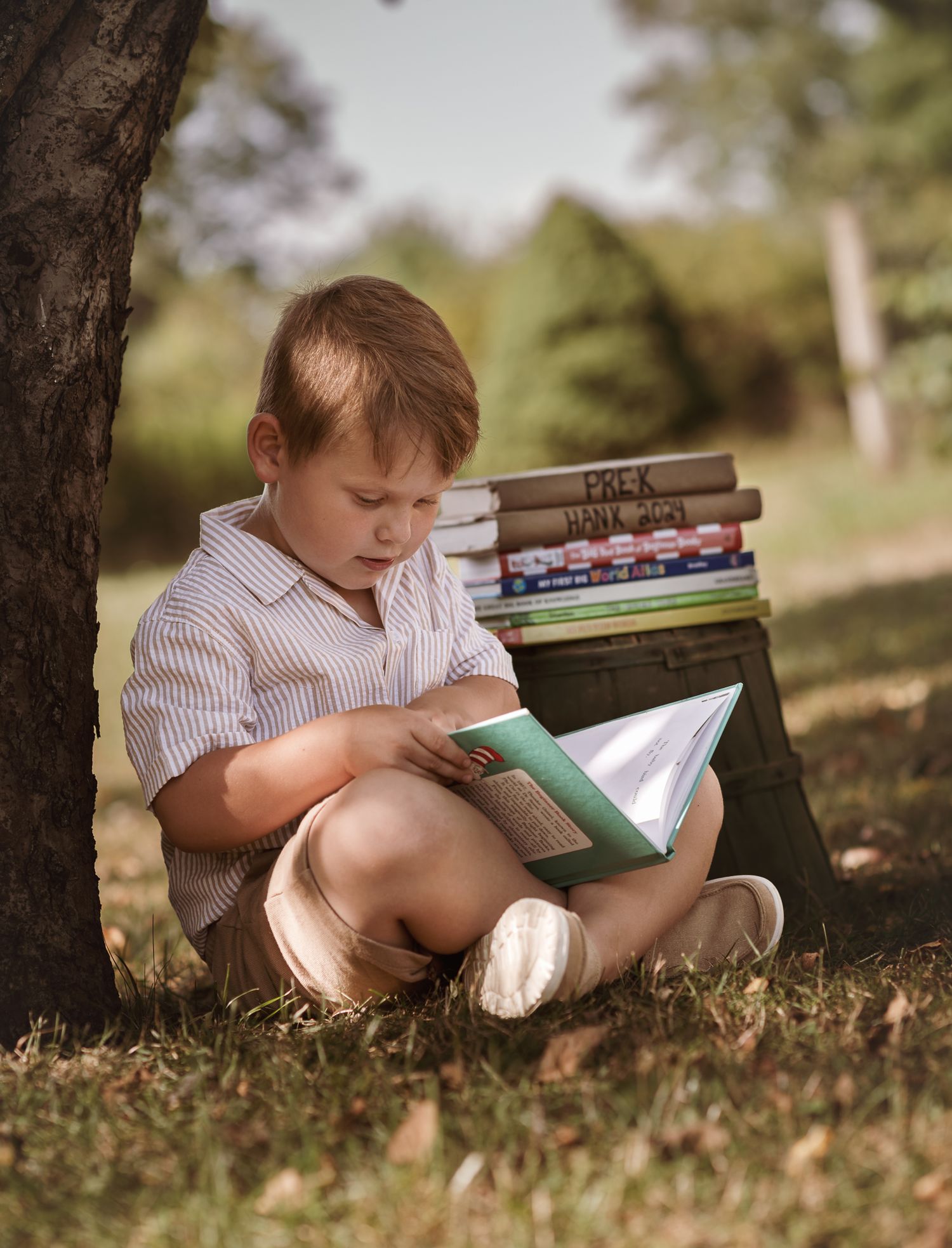 Young boy sitting under a tree reading a book outdoors, with a stack of books beside him on a sunny day.