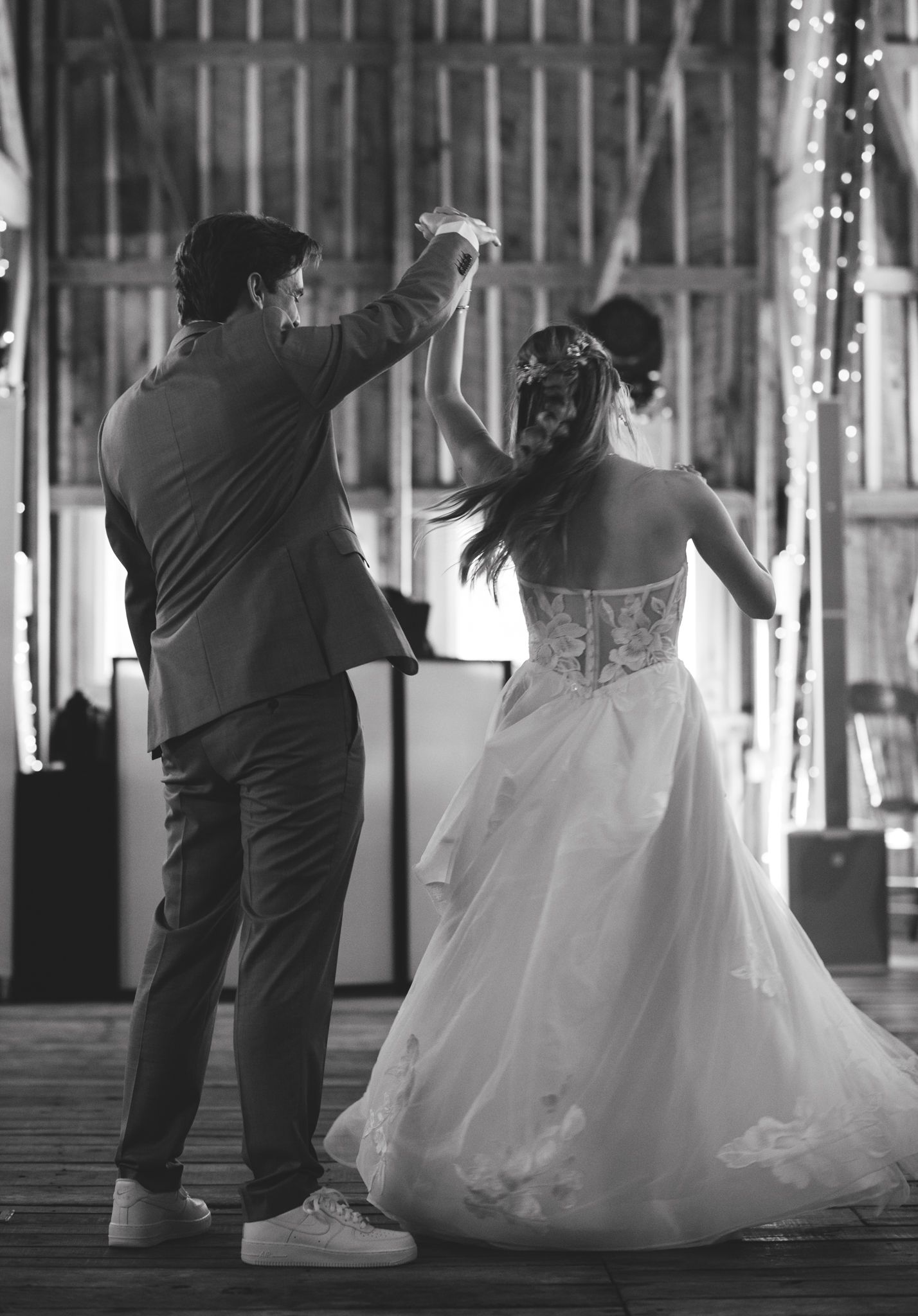 Black and white photo of bride and groom sharing first dance in a rustic barn with string lights.