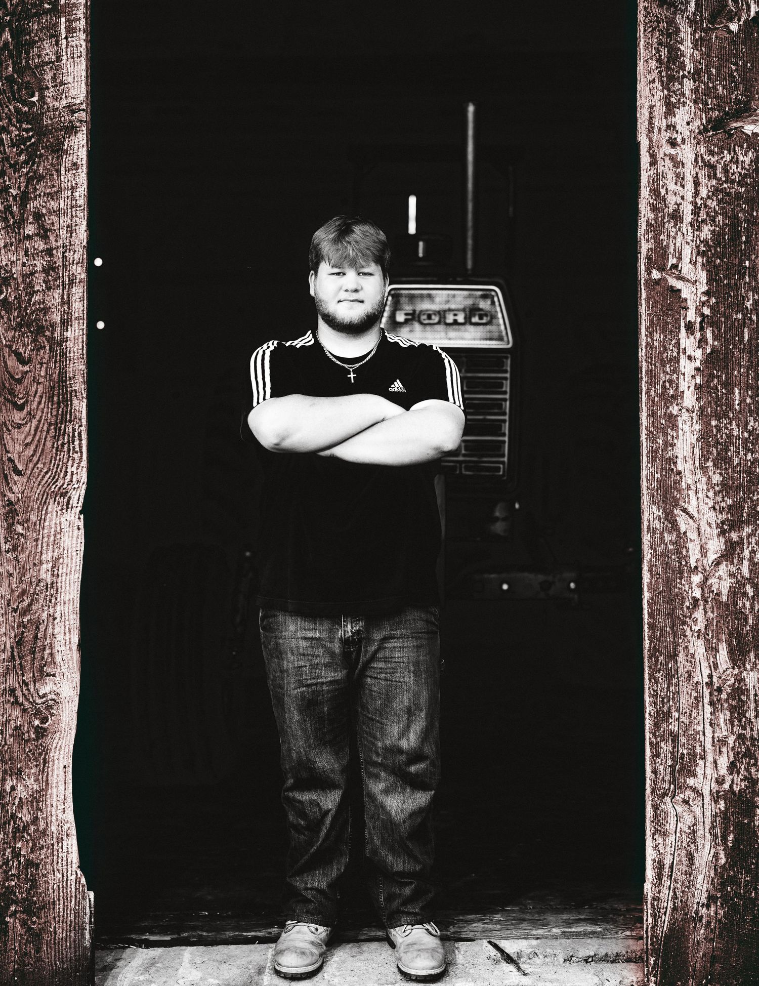 Black and white photo of a young man in black Adidas shirt, arms crossed, holding a radio, standing in a doorway.