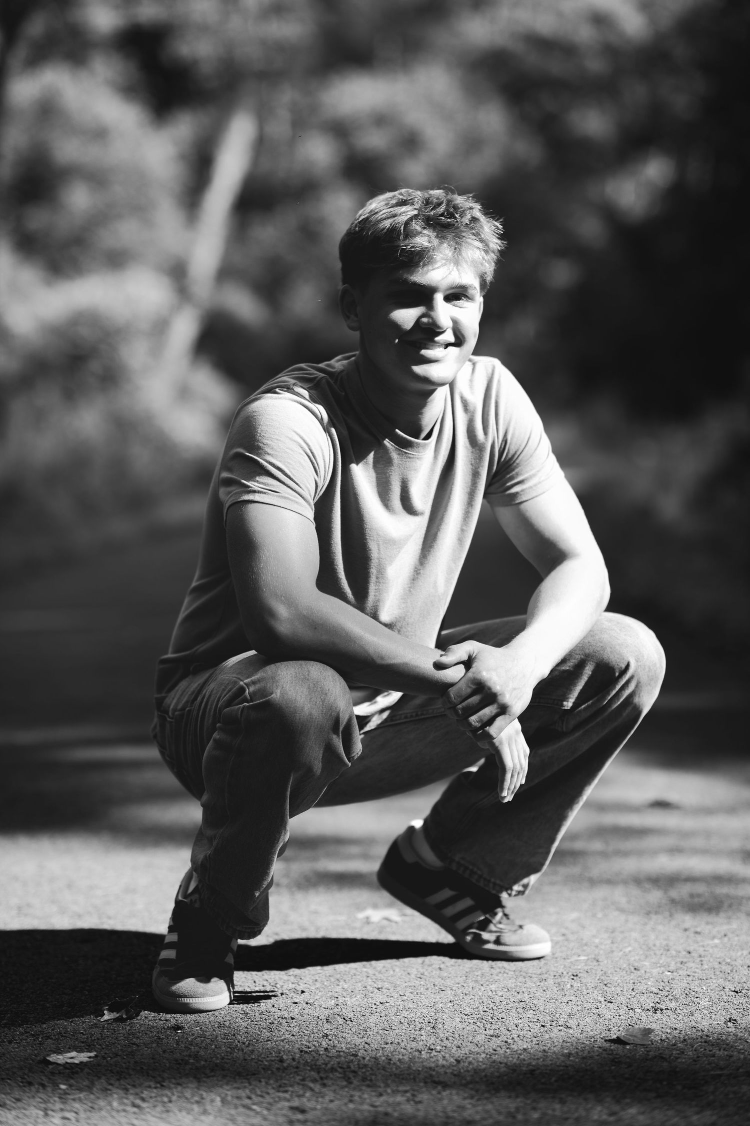Black and white photo of a smiling young man crouching outdoors in a t-shirt and jeans.