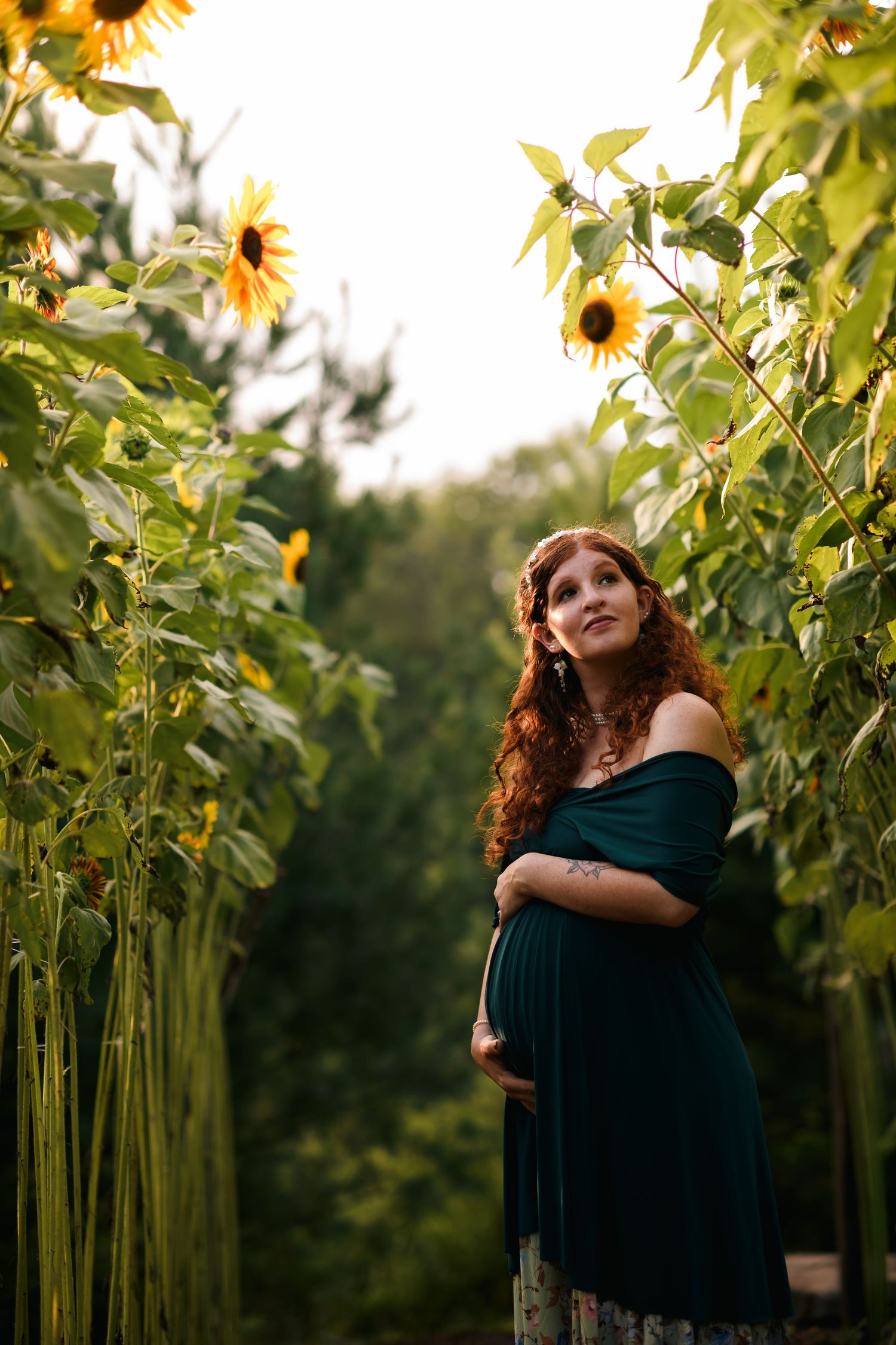Pregnant woman in teal off-shoulder dress standing among tall sunflowers, cradling her belly outdoors.