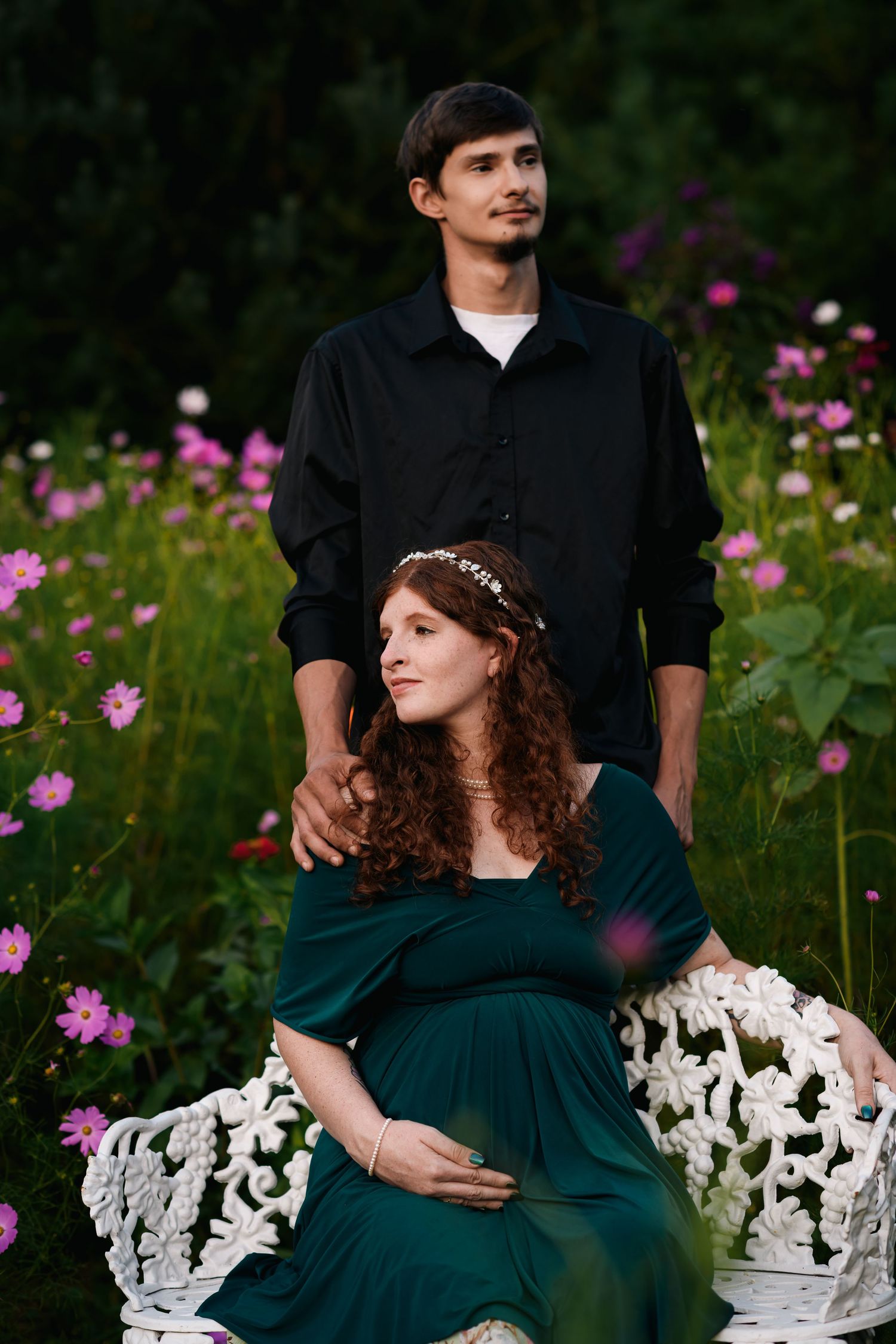 Pregnant woman in green dress sits on white bench with man in black shirt among pink flowers in garden.