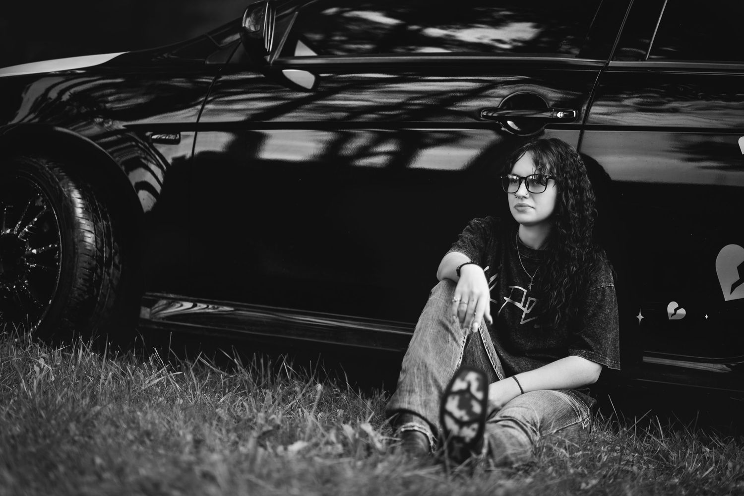 Black and white photo of a young woman with curly hair and glasses sitting on grass beside a classic car.