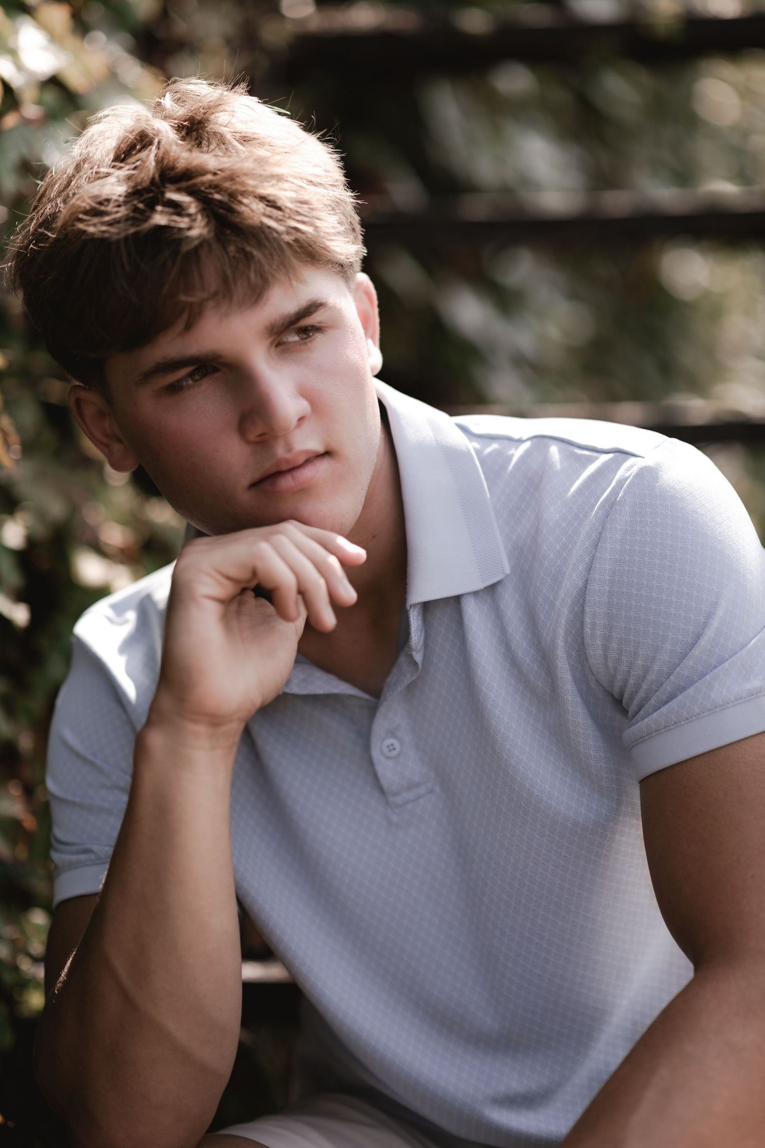 Young man in white polo shirt sitting outdoors, resting chin on hand with a thoughtful expression.