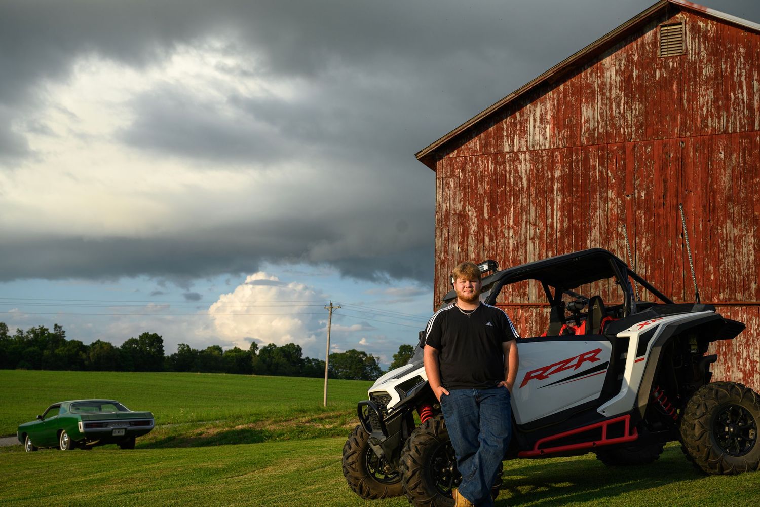 A man leans against a white Polaris RZR UTV parked beside a weathered red barn on a cloudy day.